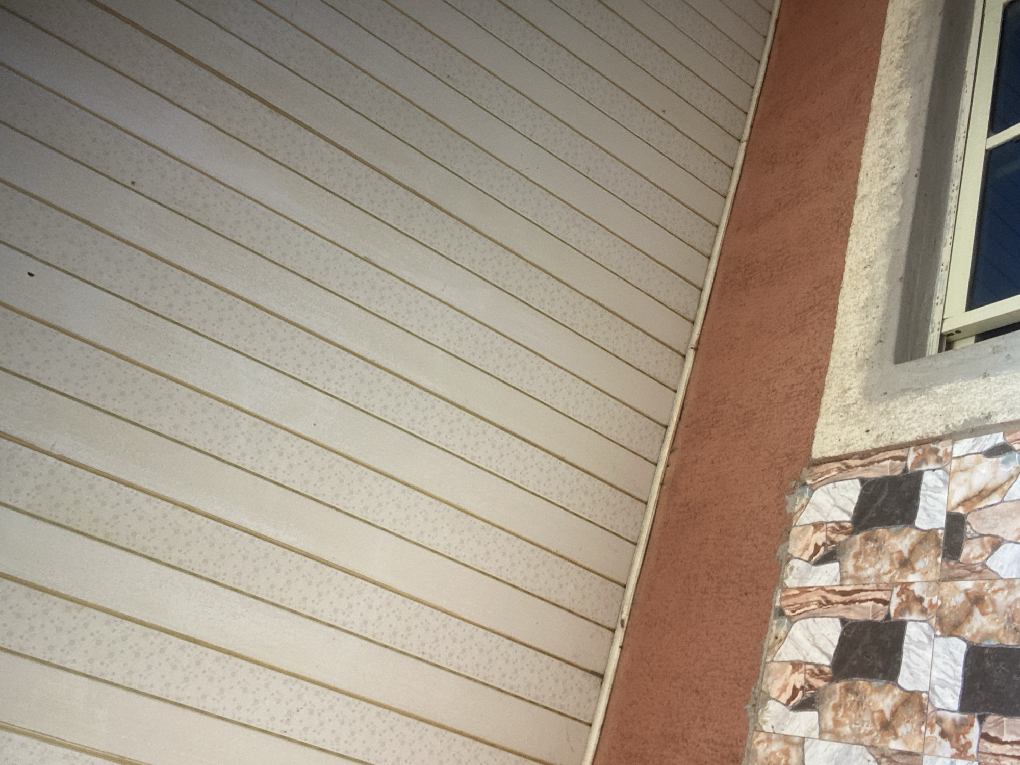 This image shows a close-up view of the exterior of a building in Jauro Yinu, Nigeria. The upper portion of the image features a white, slatted ceiling with thin, golden-colored dividers between the slats. The surface of the ceiling appears to have a speckled texture. Below the ceiling is a band of reddish-brown textured stucco. The bottom portion of the image displays a section of wall clad in square tiles with a marble-like pattern in shades of brown, beige, and black. A portion of a window frame is visible in the lower right corner. No people or animals are present in the image. The lighting suggests it is daytime.