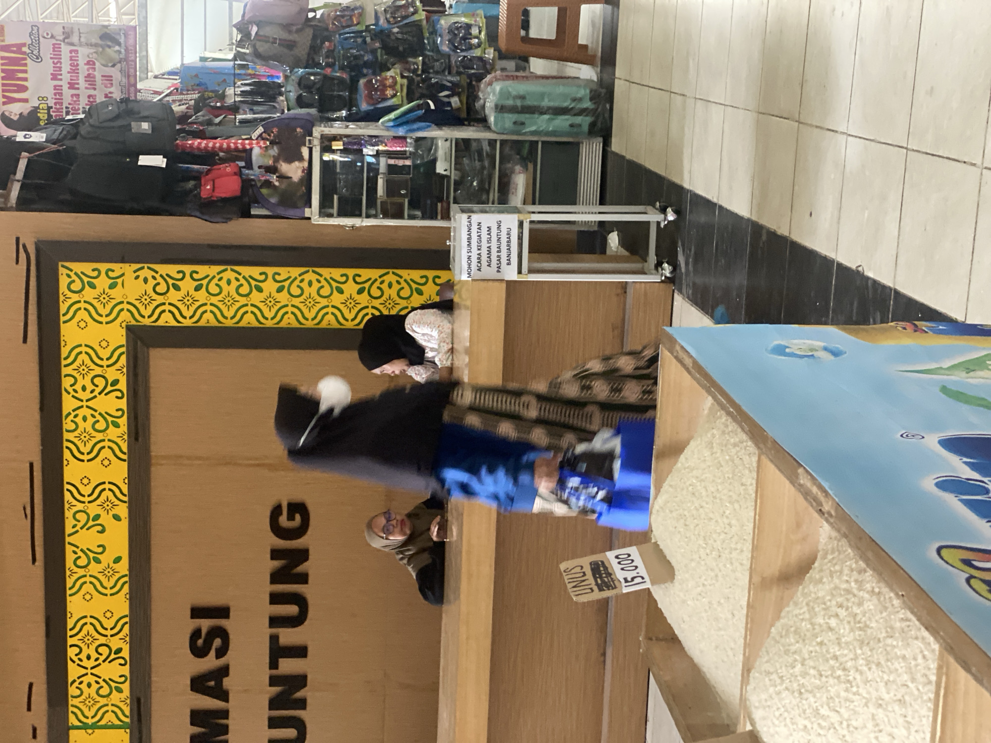 The image depicts an indoor scene, likely a market or a shop, in Banjarbaru, Indonesia. In the foreground, two wooden bins filled with white rice are visible. A blue and white sign with cartoonish figures is attached to the front of the bin on the right. To the left of the rice bins, a sign displays "LINUS 15.000".
In the mid-ground, a wooden counter is present. Two women are behind the counter. One woman, wearing glasses and a headscarf, is leaning on her arms on the counter. Another woman, wearing a black headscarf and a mask, is walking past the counter, carrying a blue plastic bag. Behind the counter, there is a sign with yellow and green decorative patterns. The text on the counter sign reads "MOHON SUMBANGAN ACARA KEGIATAN PASAR BAUNTUNG BANJARBARU".
Further in the background, to the right of the counter, a metal display shelf unit is filled with various items, including bags, backpacks, and what appear to be toiletries or personal care products. Bags are also hanging from above. A turquoise suitcase stands next to the shelf unit. The floor is tiled with cream-colored squares. The overall lighting suggests an indoor environment with artificial lighting.
