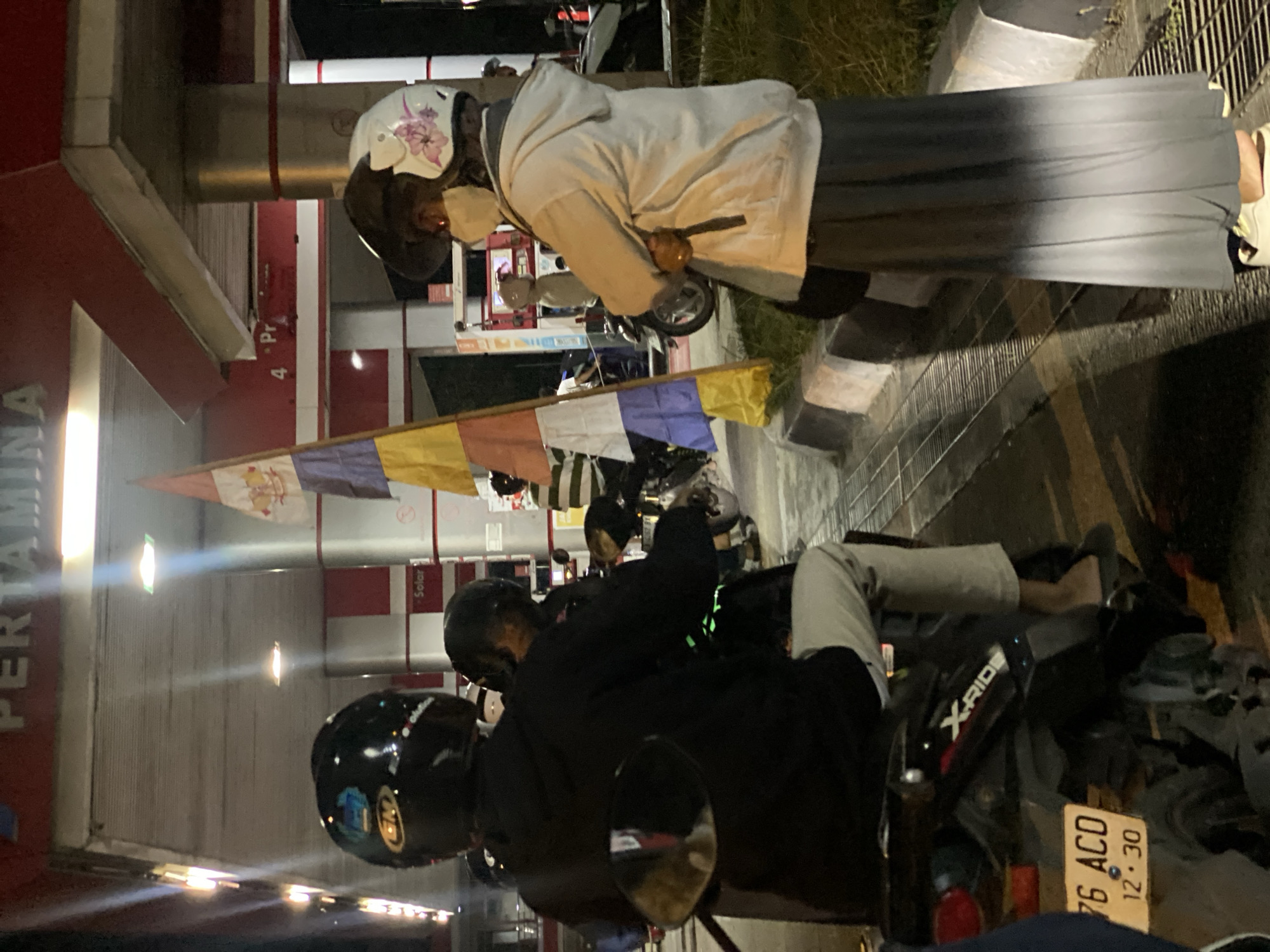 This is a medium shot, eye-level, medium-format photograph taken at night at a Pertamina gas station in Gambut Barat, Indonesia. The scene depicts several people on motorcycles. In the foreground, from left to right, a person wearing a black helmet and a black hooded jacket is seated on a motorcycle, facing away from the camera. To their right, a person wearing a black helmet is partially visible, also on a motorcycle. To the right of these individuals, a person wearing a white helmet with a floral design and a light-colored hooded jacket over a long grey skirt stands with their back to the camera, holding a cell phone. Behind them, a gas pump is visible. A flag with horizontal stripes of white, red, and yellow is prominently displayed on a pole to the left of the standing individual. The ground is paved, and a drain grate is visible in the lower right portion of the image. The overall lighting is artificial, typical of a nighttime urban environment.