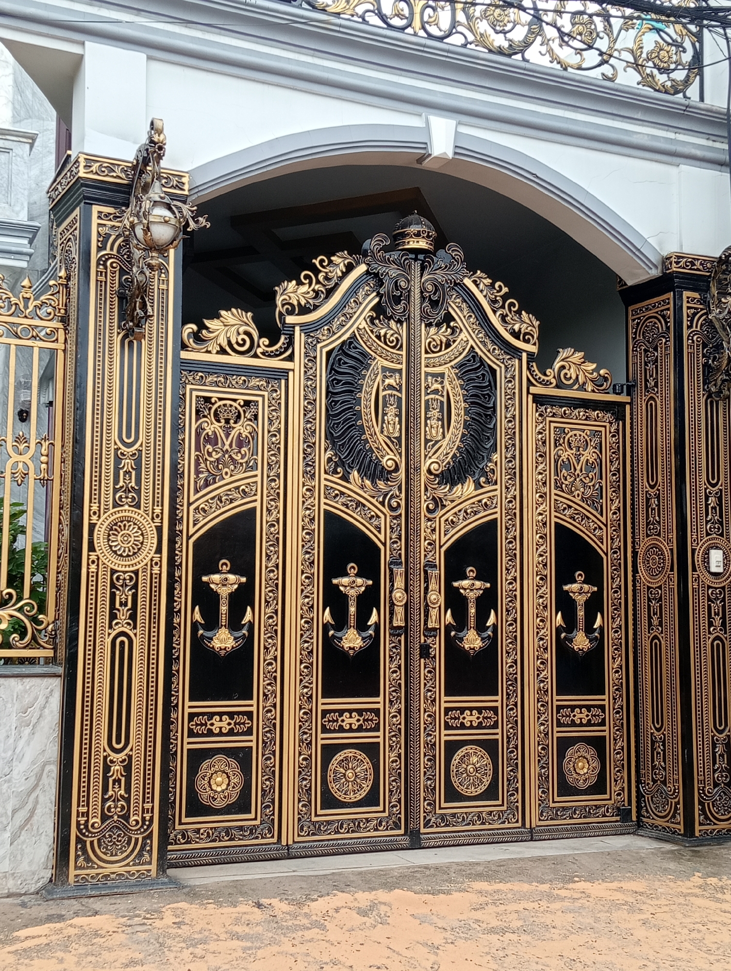 A full-length outdoor shot captures an ornate black and gold gate in front of a house. The gate is composed of four sections, with the two center sections slightly taller than the outer ones. Each section features intricate gold designs on a black background, including stylized anchors. The central sections also have a circular emblem at the top. The gate posts are adorned with similar gold detailing. A lantern-style light fixture is mounted on the gatepost on the left. The gate is beneath an arched, white doorway. The ground in front of the gate is paved and weathered. The location is Unknown, Unknown.