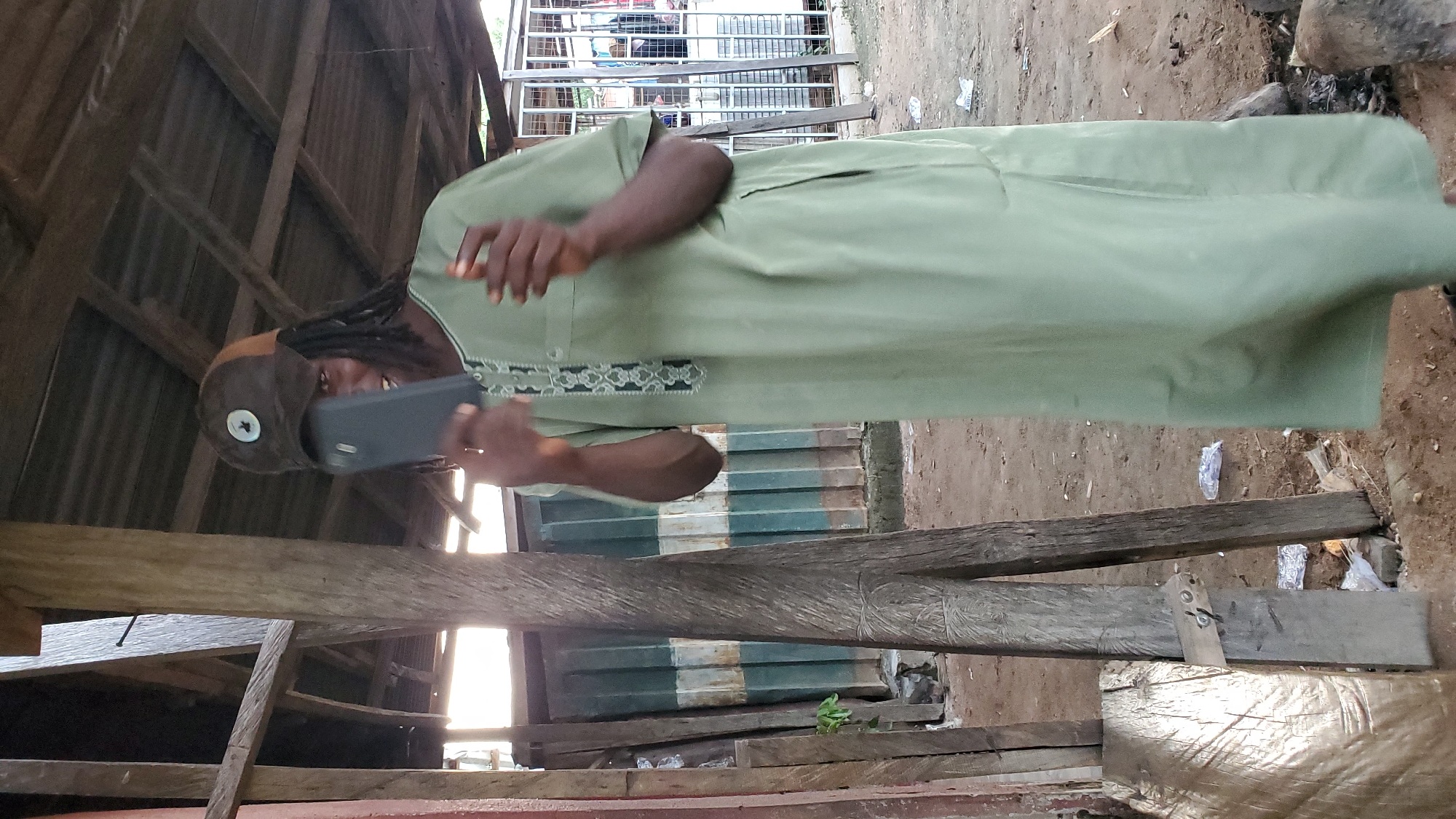 A Black male wearing a light green dashiki and a brown baseball cap is standing outdoors, holding a black smartphone in his right hand, appearing to take a selfie. The background shows a rustic, open-air structure with a corrugated metal roof supported by wooden beams and posts. Visible in the background are corrugated metal siding and a metal cage or enclosure. The ground is dirt and debris, with scattered pieces of trash. The scene appears to be in a less developed area.