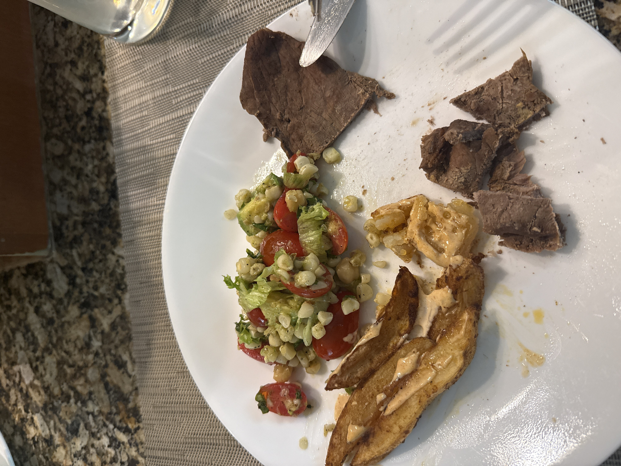 The image displays a close-up, slightly overhead view of a white plate filled with a meal, likely taken in San Bernardino Tlaxcalancingo, Mexico. The plate holds several food items: slices of cooked, brown beef, a salad, and potato wedges.
The beef appears to be a roast or similar cut, presented in irregularly shaped pieces. One larger piece is visible at the top left, with a knife blade resting on it, suggesting it's in the process of being cut. Further right, smaller, cut pieces of beef are scattered on the plate.
The salad, situated to the left of the plate, consists primarily of chopped green lettuce, halved red cherry tomatoes, and numerous yellow corn kernels. Some of the tomatoes are visible at the bottom left, slightly separated from the main salad pile.
At the bottom right, several golden-brown potato wedges are arranged, covered in a light-colored, creamy sauce or dressing. Near these, there are also some smaller, saucy bits which might be more of the potato or another ingredient.
A metal fork with distinct tines is visible towards the top right, partially on the plate, and a metal knife is positioned near the top of the large beef slice. The meal is served on a textured placemat, possibly made of woven fabric, which rests on a speckled granite or stone countertop. A partial view of a clear glass is seen at the top left corner. No people are visible, and there is no visible text in the image. The setting suggests an indoor dining experience, possibly lunch or dinner.