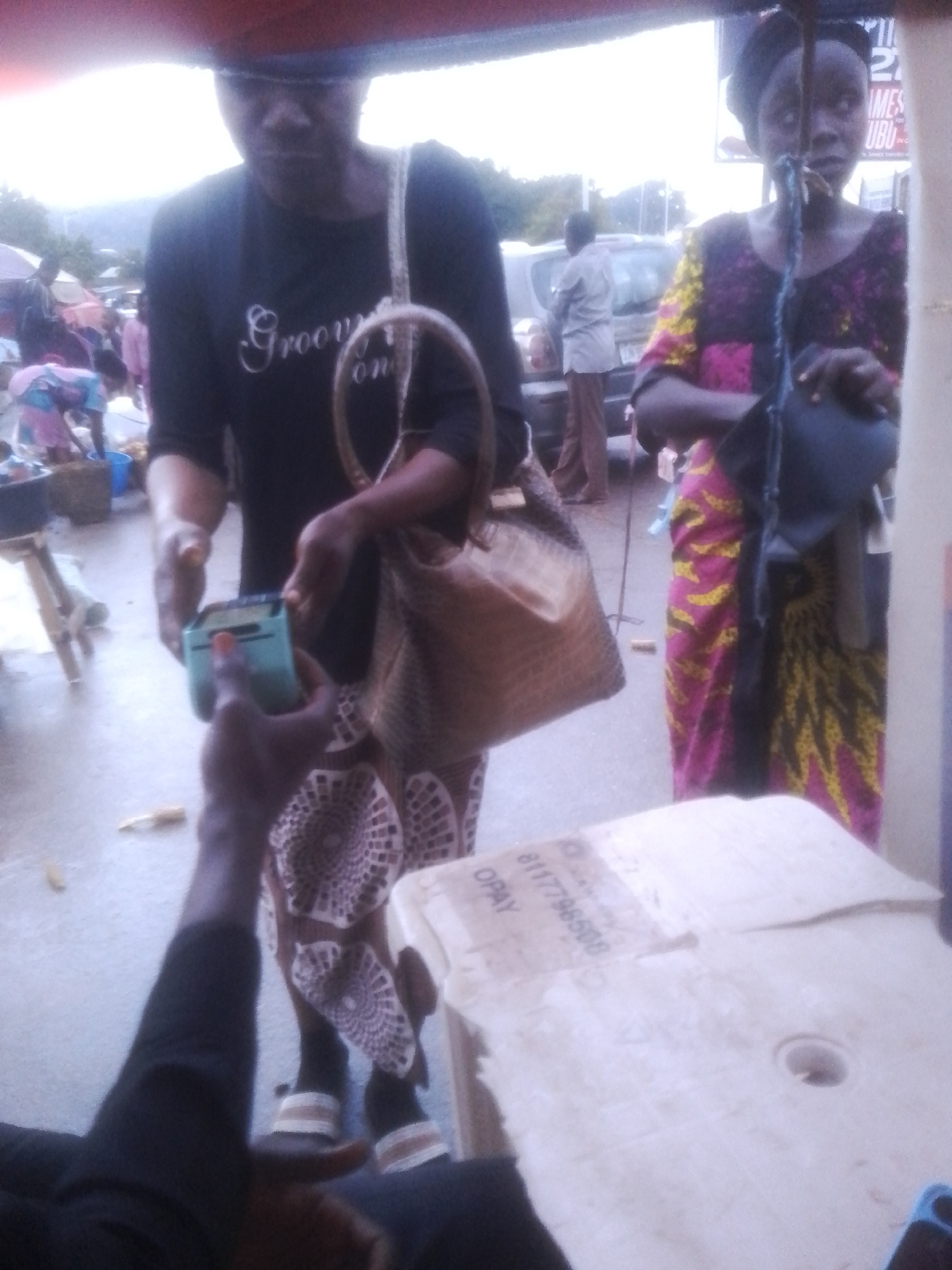 The image depicts an outdoor scene in Gwong, Nigeria, likely a market or busy street. In the foreground, a person's arm and hand are extended, holding a turquoise mobile payment device. This hand is interacting with the hand of another person, who is also holding the device. The second person appears to be a woman wearing a black t-shirt with "Groovy" written on it in white script and a brown woven bag slung over her shoulder. Behind her, another woman is visible, wearing a colorful patterned dress and a dark head covering. She is holding a dark object, possibly a bag or a piece of fabric, over her shoulder.
In the background, other people and vehicles can be seen, suggesting a public space with ongoing activity. The lighting suggests it is daytime, possibly overcast or late afternoon. A sign with visible text is present in the upper right portion of the image, though the text is partially obscured and difficult to read. Another piece of paper or cardboard with text is on a surface in the lower right of the frame, displaying "OPAY" and a series of numbers "8117796508". The overall impression is one of commerce and daily life in a Nigerian setting.