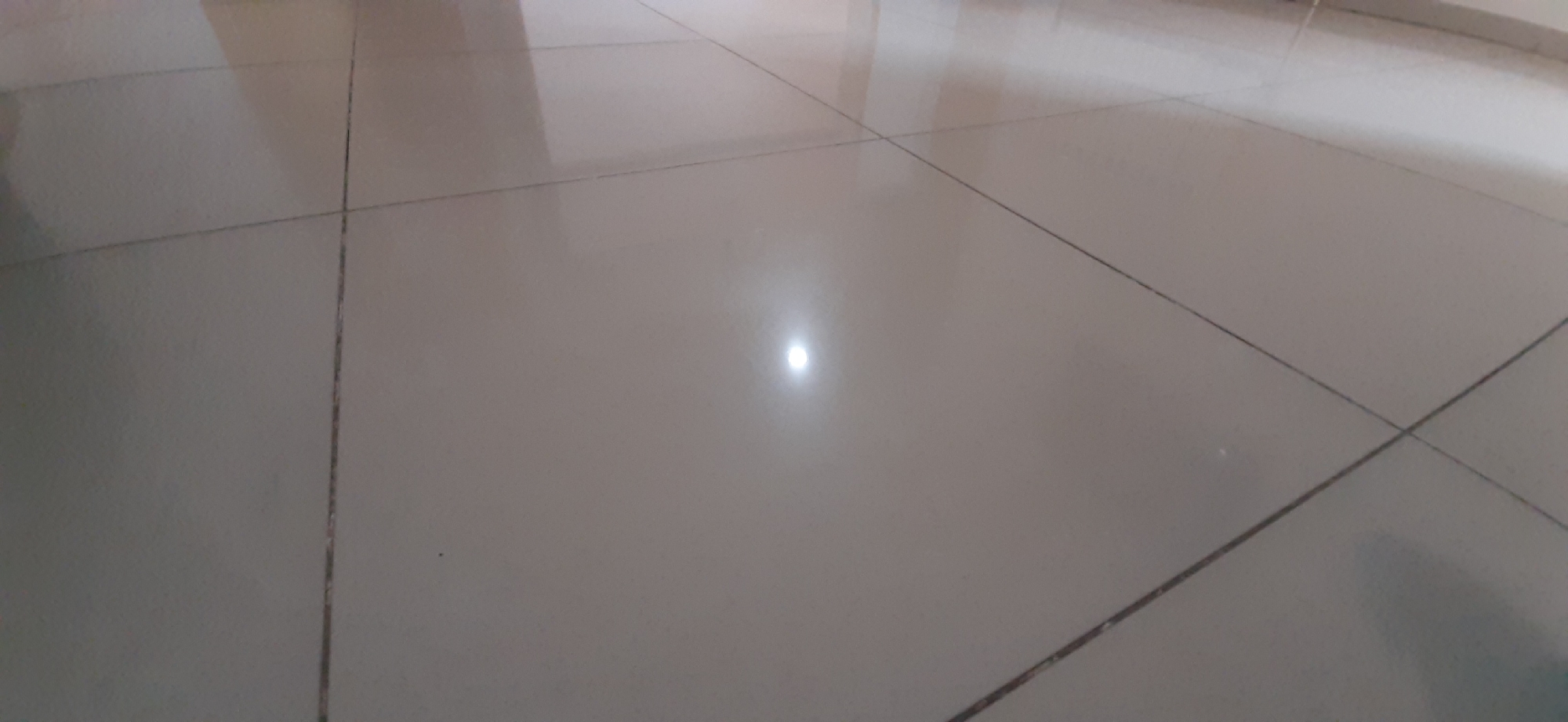 The image shows a close-up, slightly angled view of a tiled floor. The tiles are a light, neutral color, possibly beige or off-white, and are laid in a grid pattern with dark grout lines. The floor is highly reflective, with a bright, circular highlight in the center of the frame, indicating a strong light source directly above or to the side. This reflection suggests the floor is clean and polished. The rest of the floor is slightly less bright, with some muted reflections of what might be walls or overhead structures visible in the upper left portion of the image. There are no people, objects, or text visible in the frame. The scene is simple and focuses solely on the texture and reflective quality of the tiled surface. Given the location context of Yessentuki, Russia, this could be the floor of a building, such as a home, hotel, or public space. The lighting suggests it could be daytime, with artificial lights contributing to the bright reflection.