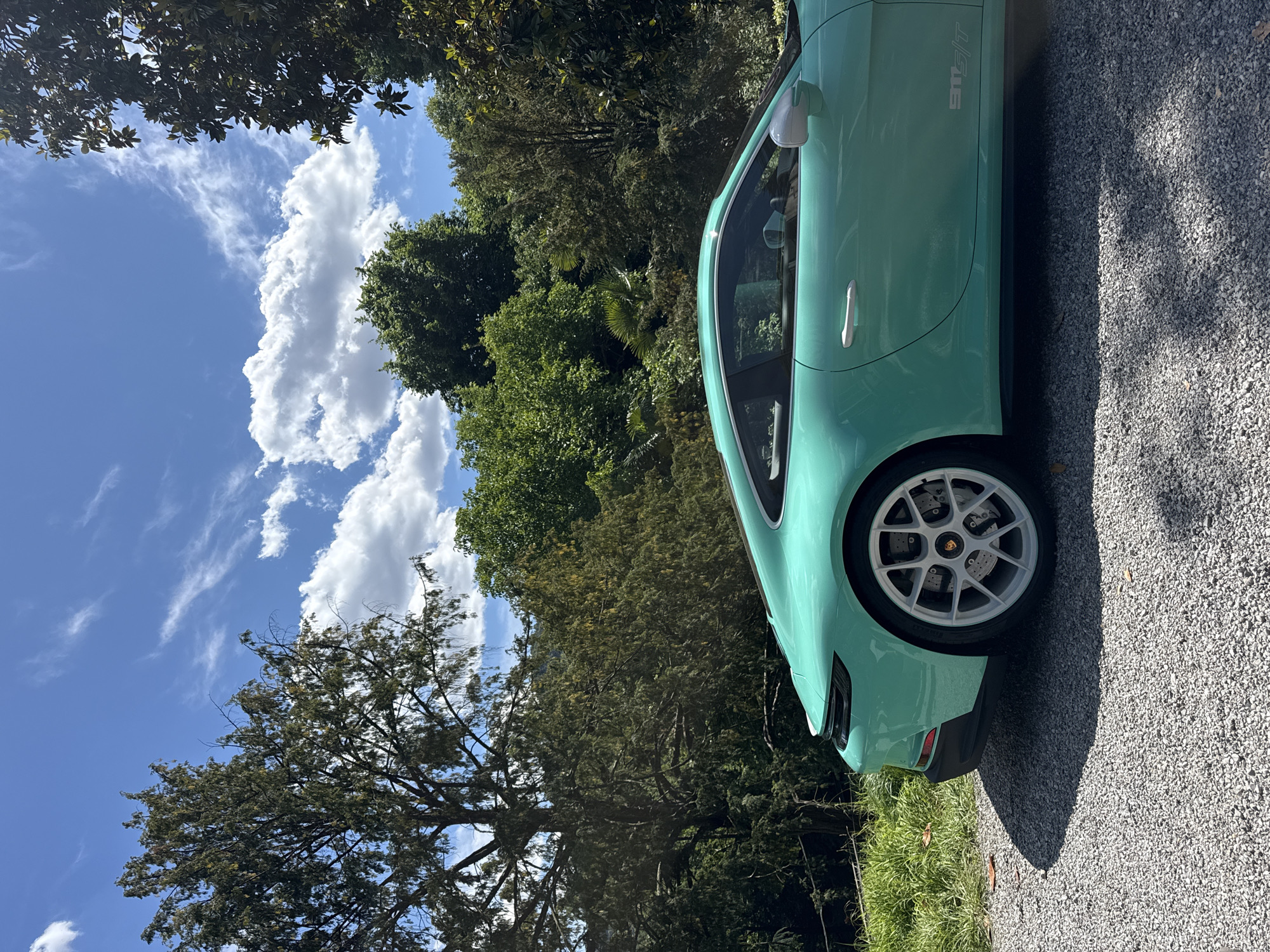 The image shows a light turquoise Porsche 911 GT3 parked on a gravel driveway. The background consists of lush green trees and a bright blue sky with scattered white clouds. The car appears to be in a residential area, possibly in the city of Como, Italy, based on the landscape and architecture. The image is a medium shot, focusing on the front quarter of the car and its surroundings. The composition suggests a sunny day with clear weather.