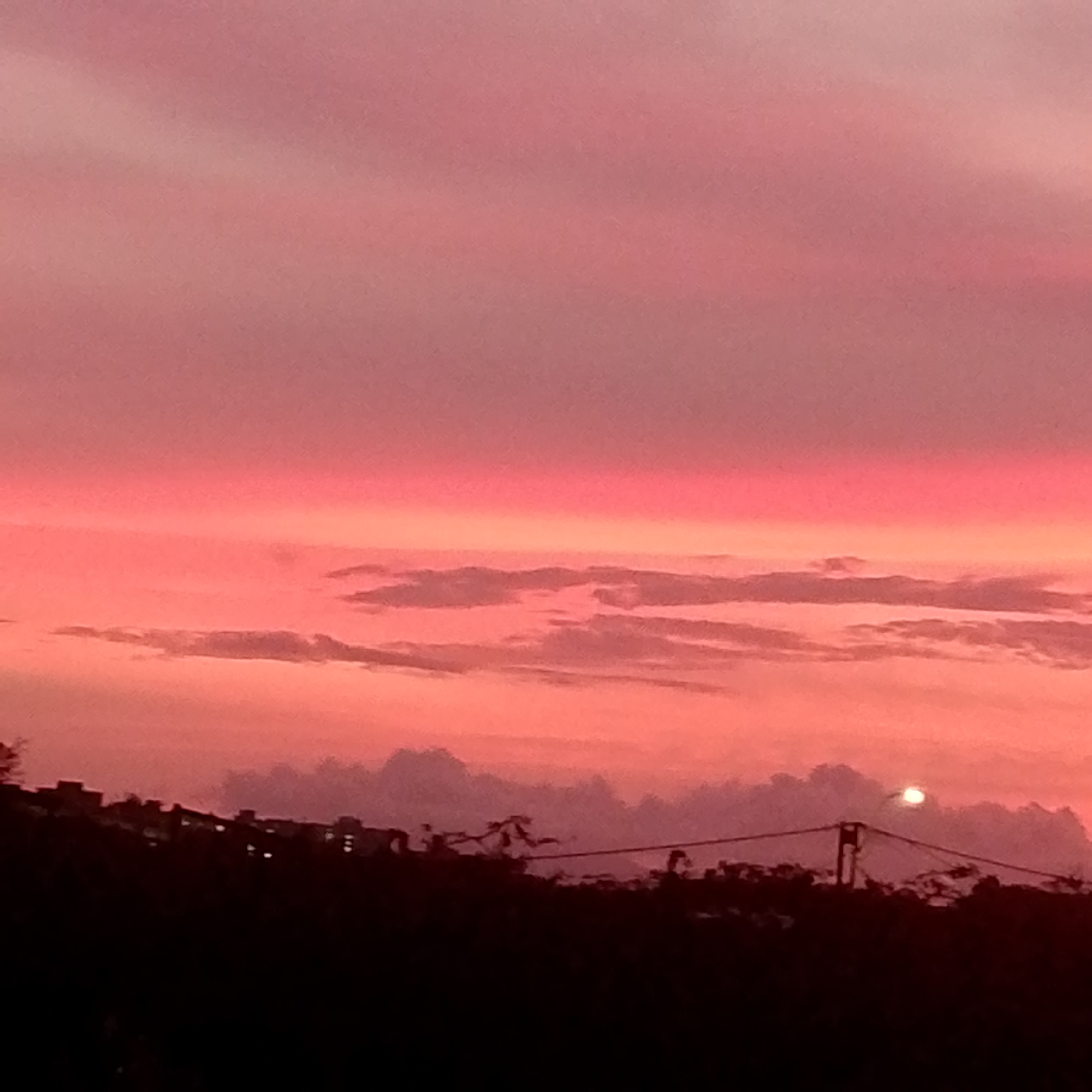 The image depicts a sunset in Altagracia, Venezuela. The sky is dominated by pink and red hues. Several layers of clouds are visible in the middle ground. The foreground features a dark, silhouetted landscape with trees and some visible structures which may be buildings. A bright light is visible in the distance, possibly the sun or a streetlight.