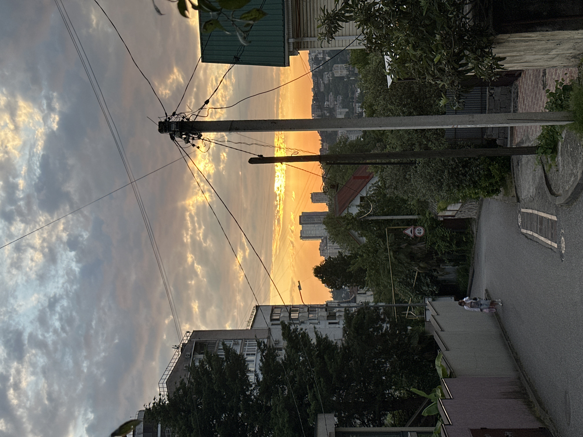 This is a photograph taken from a street in Sochi, Russia, likely during sunset. The sky is filled with dramatic clouds, with bright orange and yellow hues radiating from the setting sun, which is partially obscured by clouds. In the foreground, a street slopes downwards, bordered by a textured, light-colored wall on the left and dense green foliage and a house with a green roof on the right. A utility pole with numerous wires is prominently positioned in the mid-ground. In the distance, several tall buildings are visible, along with a cityscape that extends across rolling hills. Two people are walking down the street, appearing as small figures in the distance. The overall impression is a suburban or residential area transitioning into a more urban landscape, with the natural beauty of the sunset adding a striking element to the scene.