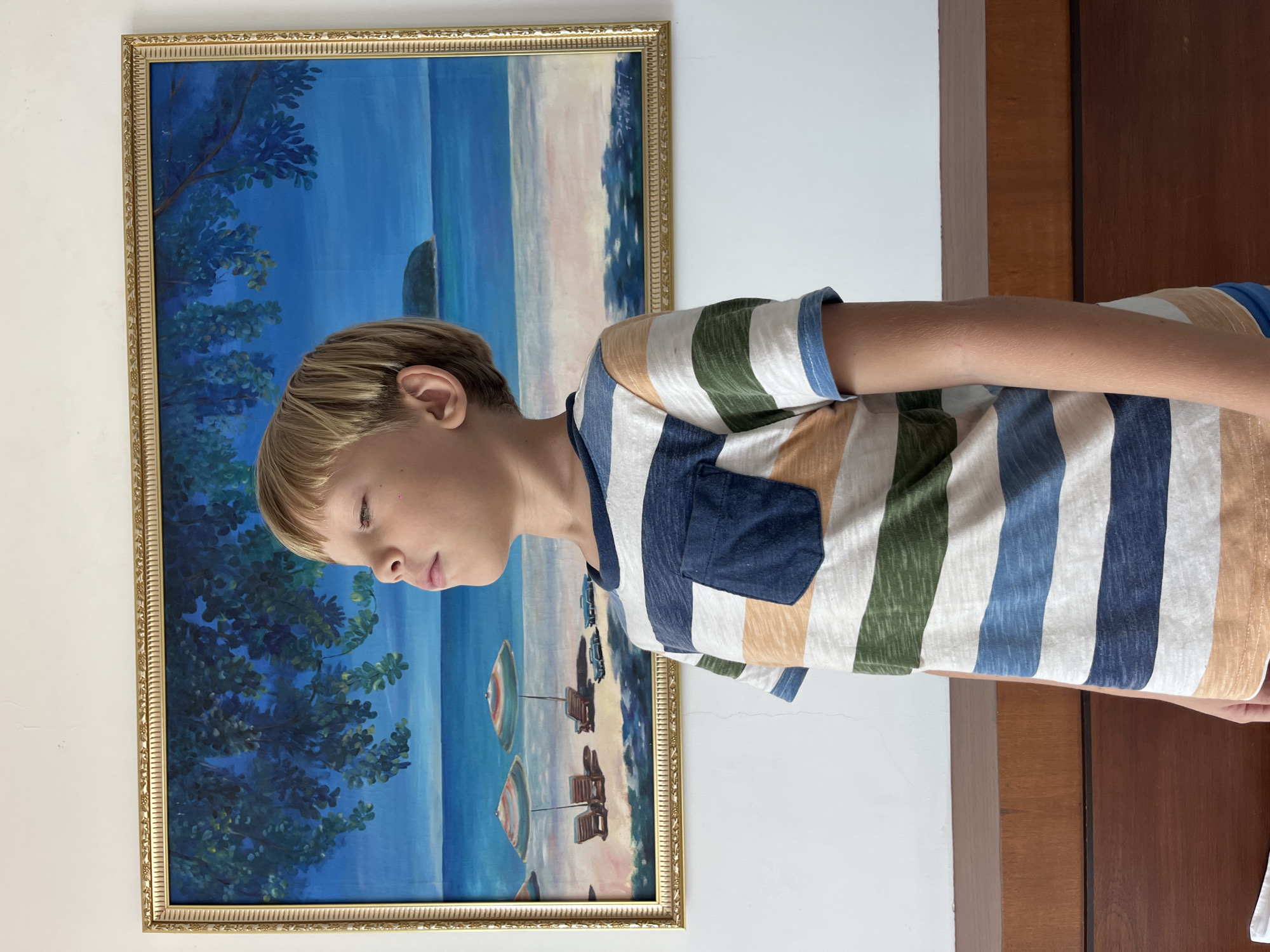 In Rawai, Thailand, a young boy with blond hair stands in front of a painting of a beach scene. The painting, enclosed in a golden frame, depicts two beach umbrellas, chairs, and a calm blue ocean under a bright sky with lush green trees in the foreground. The boy, wearing a horizontally striped t-shirt in shades of blue, green, orange, and white, has his eyes closed and is facing to the right, with his head tilted slightly upwards as if feeling the sun. He is positioned in front of a wooden cabinet. The wall behind him is plain white.