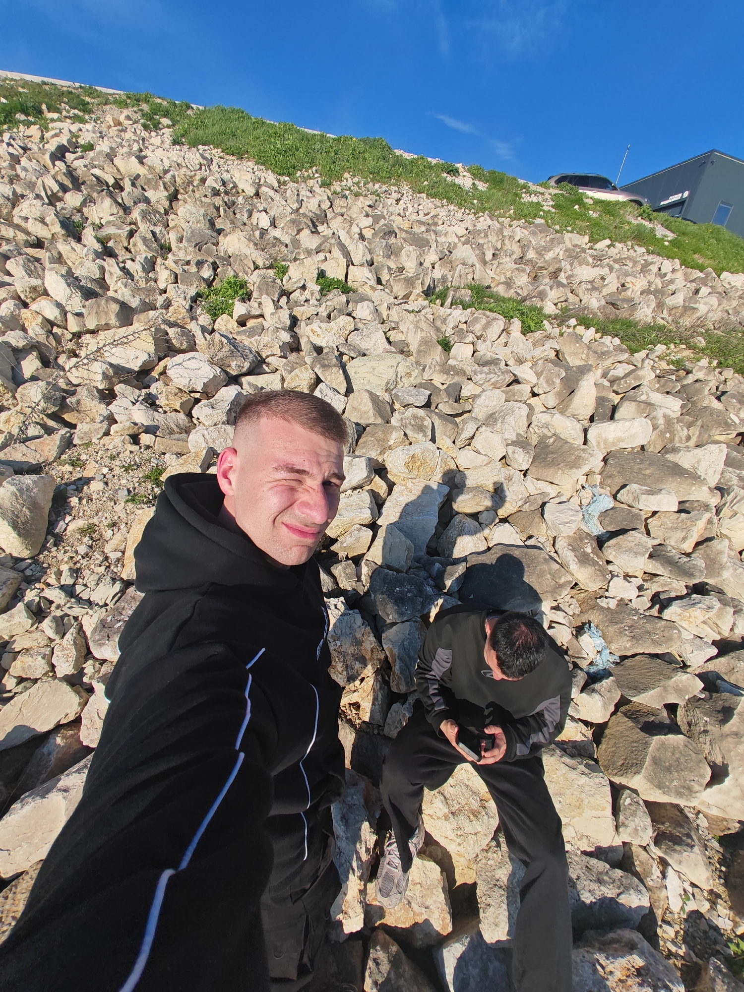 Two men stand on a rock-covered slope in Shumen, Bulgaria. The foreground shows one man in a black hoodie with white stripes, winking at the camera. Behind him, another man sits on the rocks, looking at his phone. The hillside is covered in large, light-colored rocks with patches of green grass. In the background, a modern building and a car are visible atop the slope against a clear blue sky.