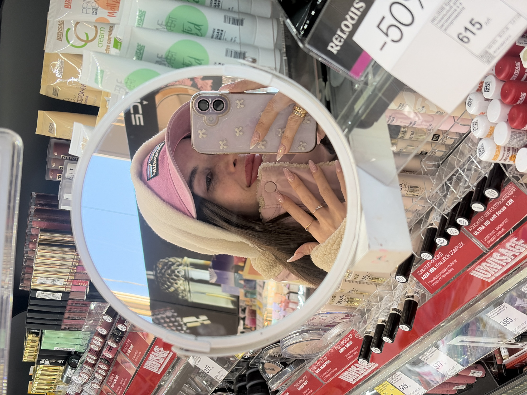 A woman is taking a selfie in a makeup mirror in a retail store. The mirror shows her face, lilac-colored phone with white flower accents, pink cap and light-colored hooded jacket. The surrounding shelves contain makeup items. Signs indicate the store is likely located in Saint Petersburg, Russia.
