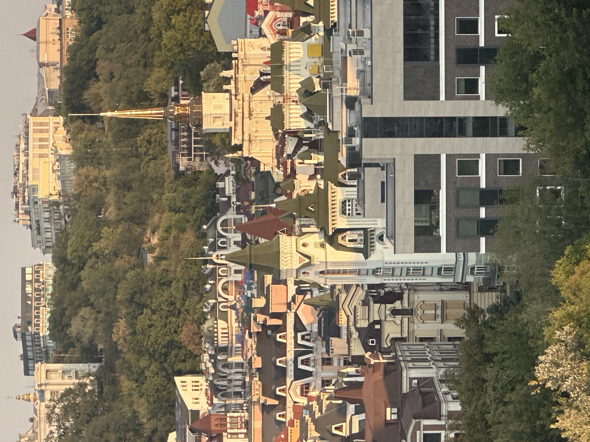 The image is an aerial view of the Podil district in Kyiv, Ukraine, featuring a dense collection of buildings with varied architectural styles. The buildings are situated on a hillside, with lush green trees interspersed throughout the scene. Dominating the foreground is a modern, multi-story building with dark brick and concrete accents, featuring numerous windows. Behind it, a cluster of older, ornate buildings with pitched roofs, dormer windows, and decorative facades are visible. Some of these older buildings have distinctive green-tiled roofs. Further up the hill, grander structures with neoclassical and baroque influences are present, some topped with spires or domes. The sky is a pale, hazy color, suggesting it is either early morning or late afternoon. The lighting casts warm tones on the buildings, highlighting their architectural details. No people are visible in the image.