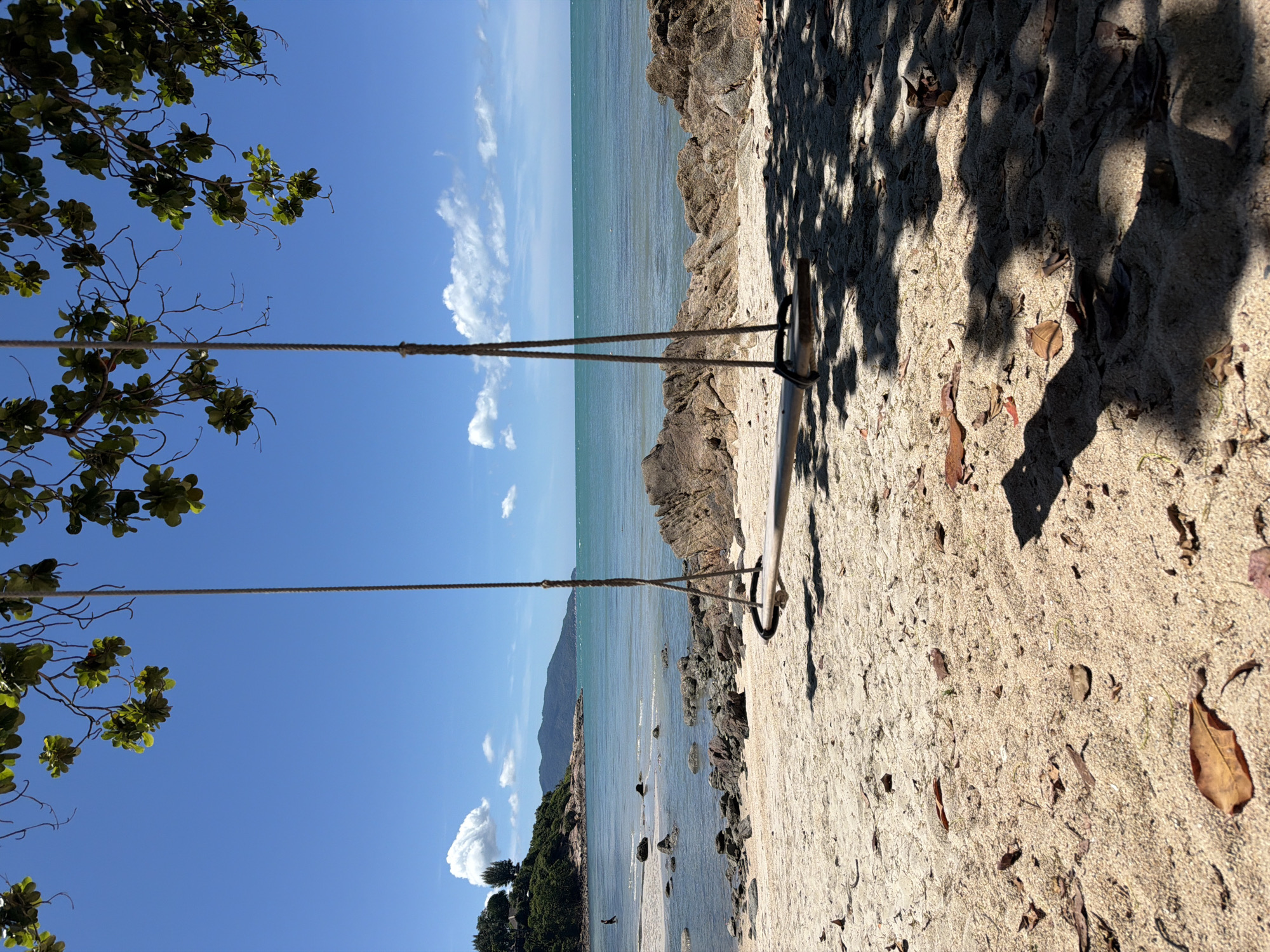 A tranquil beach scene unfolds under a bright blue sky. The turquoise water gently laps at a sandy shore dotted with rocks and fallen leaves. Lush green foliage from overhanging trees casts dappled shadows on the sand, creating a natural, serene atmosphere. The image features a rustic swing, suspended by thick ropes attached to a sturdy pole. The swing, a simple wooden plank, hints at leisurely moments and relaxation. A sense of peace and quiet pervades the scene, suggesting a secluded paradise. The time of day appears to be late morning or early afternoon, given the strong sunlight and distinct shadows. The location is identified as Baan Plai Laem, Thailand, which evokes images of tropical beauty and coastal charm. No people are visible, emphasizing the solitude of the setting. The overall impression is one of natural beauty, simplicity, and serenity, perfect for unwinding and enjoying the coastal environment.