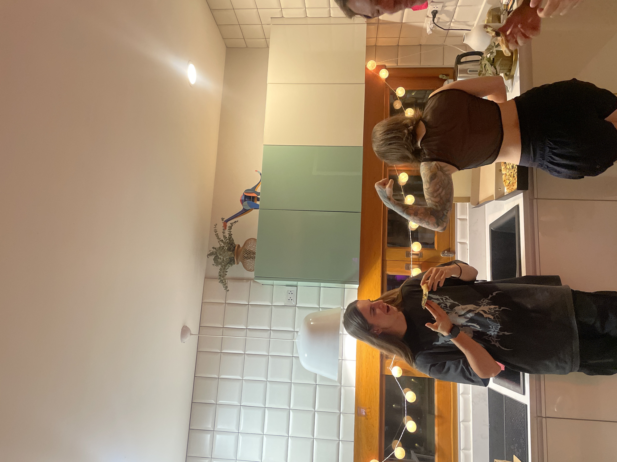 The image captures a casual evening gathering in a modern kitchen, likely located in Chang Phueak, Thailand. Three individuals are present, engaged in food-related activities.
In the foreground, a person with long, dark hair, wearing a dark t-shirt with a prominent light-colored graphic design, holds a piece of food, possibly a snack, and looks down at it. Their left wrist sports a watch. Further into the kitchen, another person with dark, shoulder-length hair and tattooed arms is seen from behind. They are wearing a sheer dark top and black shorts, standing at a kitchen counter, reaching towards or interacting with items on a tray filled with food. The head of a third person is partially visible at the top of the frame, looking towards the right.
The kitchen features white square tiled walls and light-colored cabinetry, some sections appearing in a pale green. A wooden door or window frame is decorated with a string of warm white globe lights, contributing to a cozy, evening ambiance. On the counter, a white teapot, a small potted plant, and a tray of food are visible. A colorful, abstract, bird-like wall decoration adds a unique artistic touch to the wall. Recessed ceiling lights provide additional illumination, confirming an indoor setting during the evening or night. The overall scene suggests a relaxed and social occasion focused on food. No discernible text is visible.