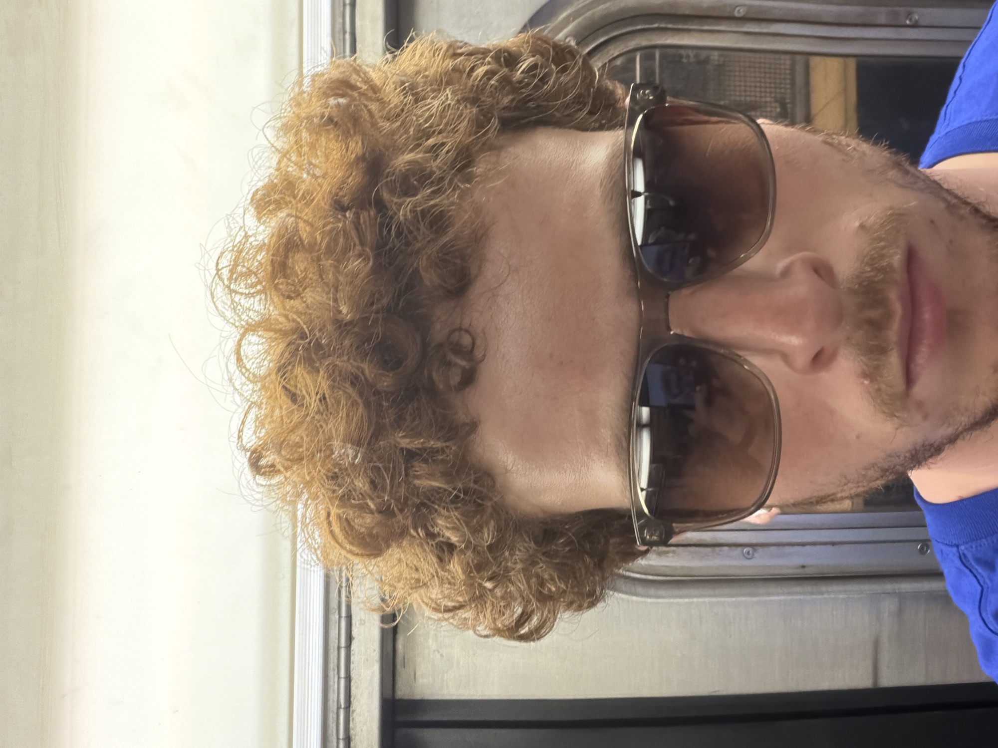 A man with curly brown hair, sunglasses, and a mustache is standing on a subway train in New York City. The background shows the interior of the subway car. He is wearing a blue shirt.