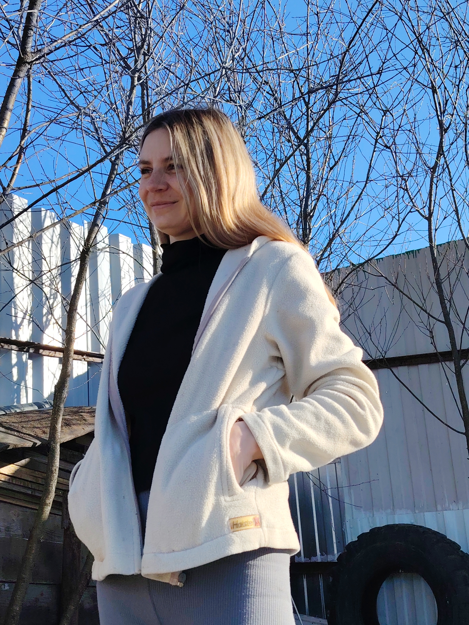 A woman stands in a backyard or industrial area, wearing a black turtleneck sweater, grey pants, and an off-white fleece jacket with her hand in one pocket. A pile of lumber sits to her left, and a large tire is visible to her right. A corrugated metal fence forms the backdrop, with leafless trees silhouetted against a clear blue sky. The location is Chaikovsky, Russia.