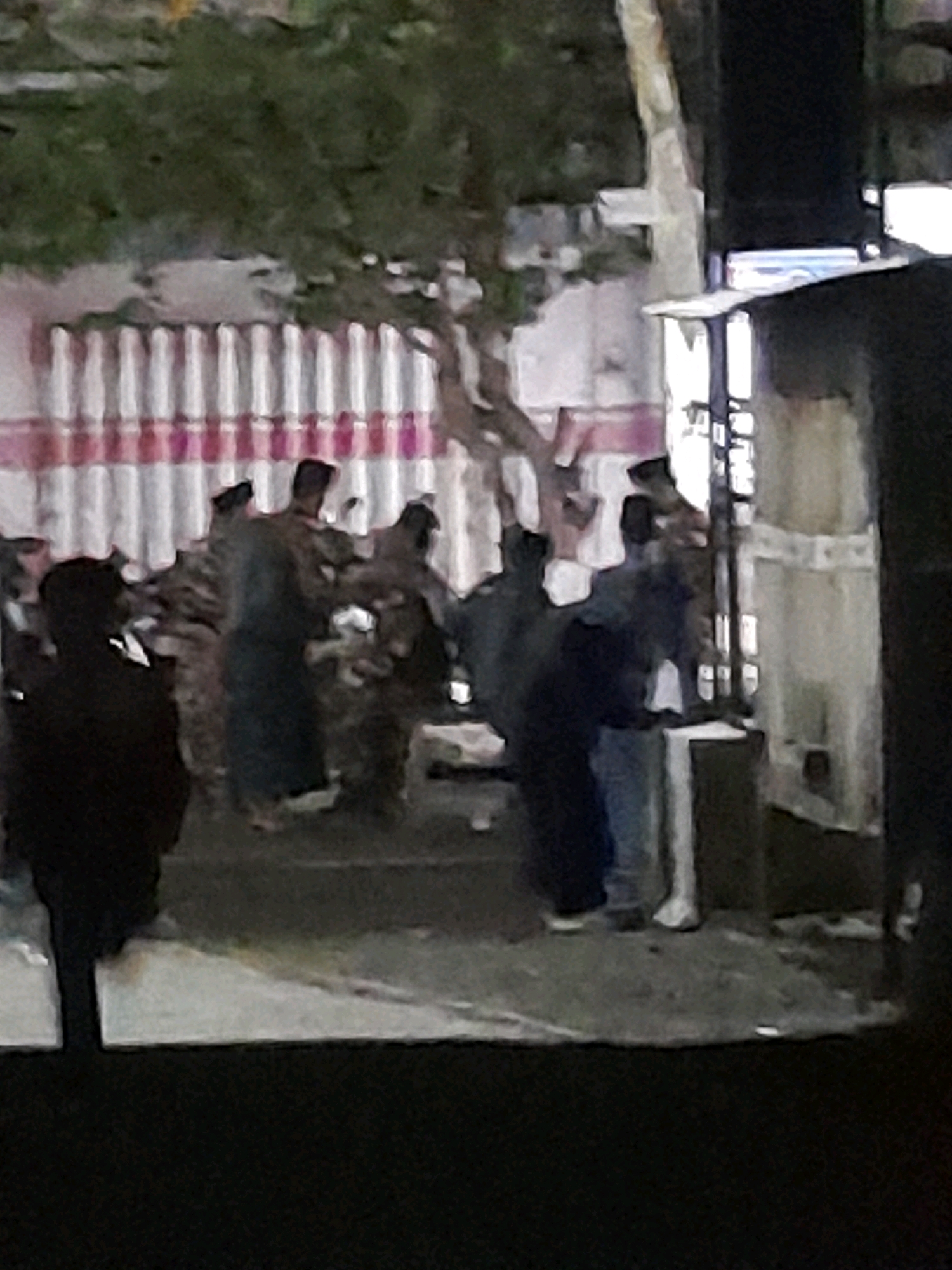The image shows a dimly lit outdoor scene with several individuals. In the foreground left, a person is silhouetted in dark clothing. Behind them, a group of people are gathered. One person in the center appears to be wearing a long, dark blue garment. To their right, another individual is wearing light blue pants. The background features a fence with vertical white bars and pink horizontal accents, possibly a wall or structure. There are trees with dark foliage above the fence. A large, dark structure is visible on the right side of the image, obscuring part of the background. The ground appears to be rough, paved, or dirt. The overall impression is of a clandestine or informal gathering.
