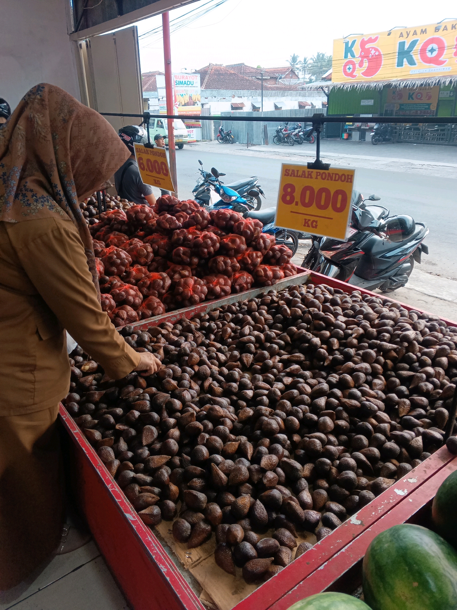 The image depicts an outdoor market scene in Garut, Indonesia, likely a fruit stall. In the foreground, a woman wearing a brown hijab and a brown uniform is seen selecting salak pondoh (snake fruit) from a large red bin. Two separate piles of salak pondoh are visible, one in netting and another in a bin, with prices displayed on yellow signs: "SALAK PONDOH 8.000 KG" and another sign partially obscured that reads "6.000 KG". In the background, motorcycles are parked, and buildings with signage are visible, including a bright yellow sign with red lettering that appears to be for a store or restaurant. The environment suggests a busy commercial area, possibly a roadside market.