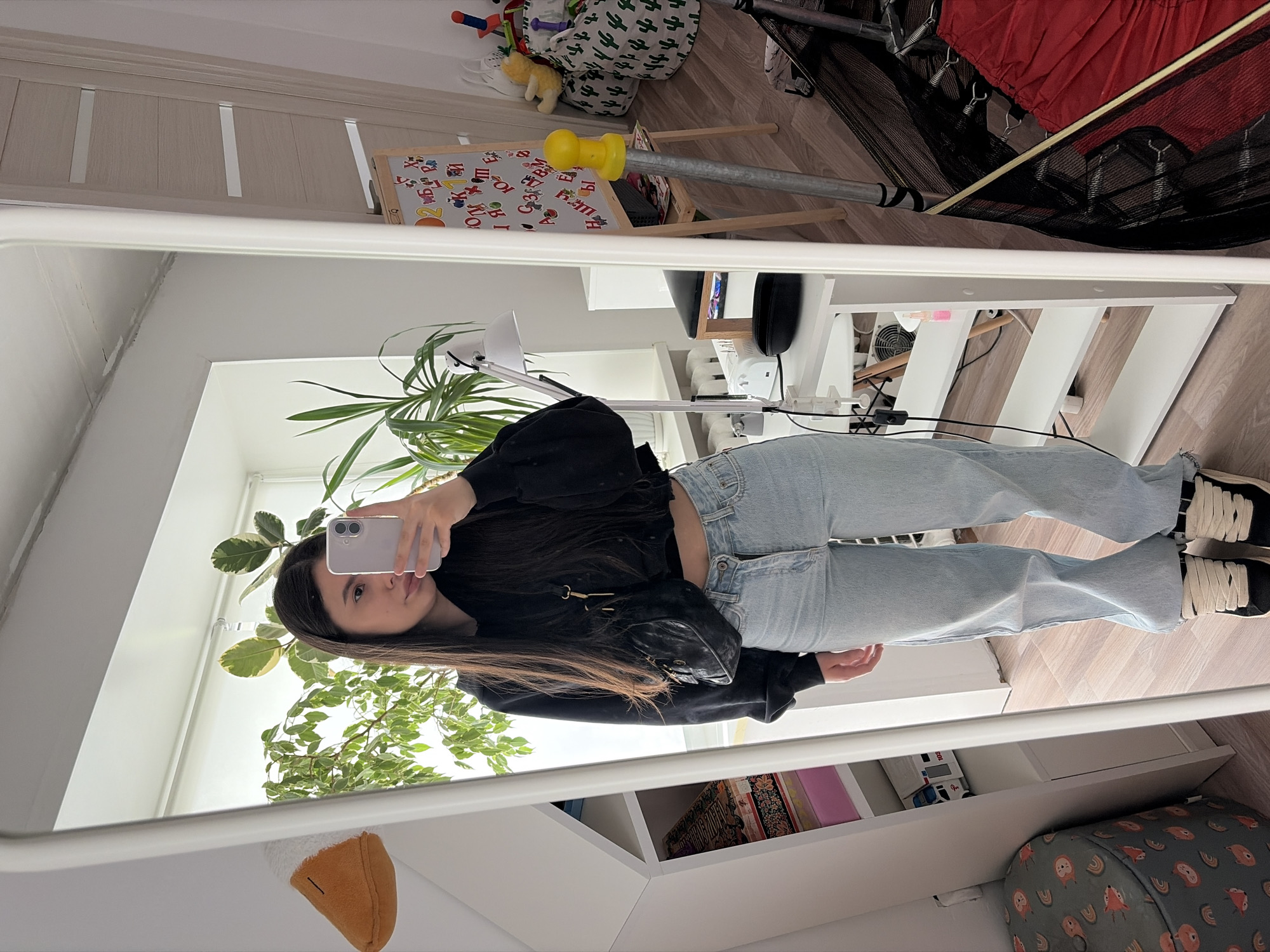 A person is taking a mirror selfie indoors. The location appears to be a room in Chaikovsky, Russia. The person is holding a smartphone and facing the mirror, wearing a black jacket, light blue jeans, and black and white sneakers. The background includes a white door, a child's easel, a trampoline, a desk with electronics, a houseplant, and a white shelving unit. A stuffed animal is visible in the bottom left corner of the mirror's reflection.