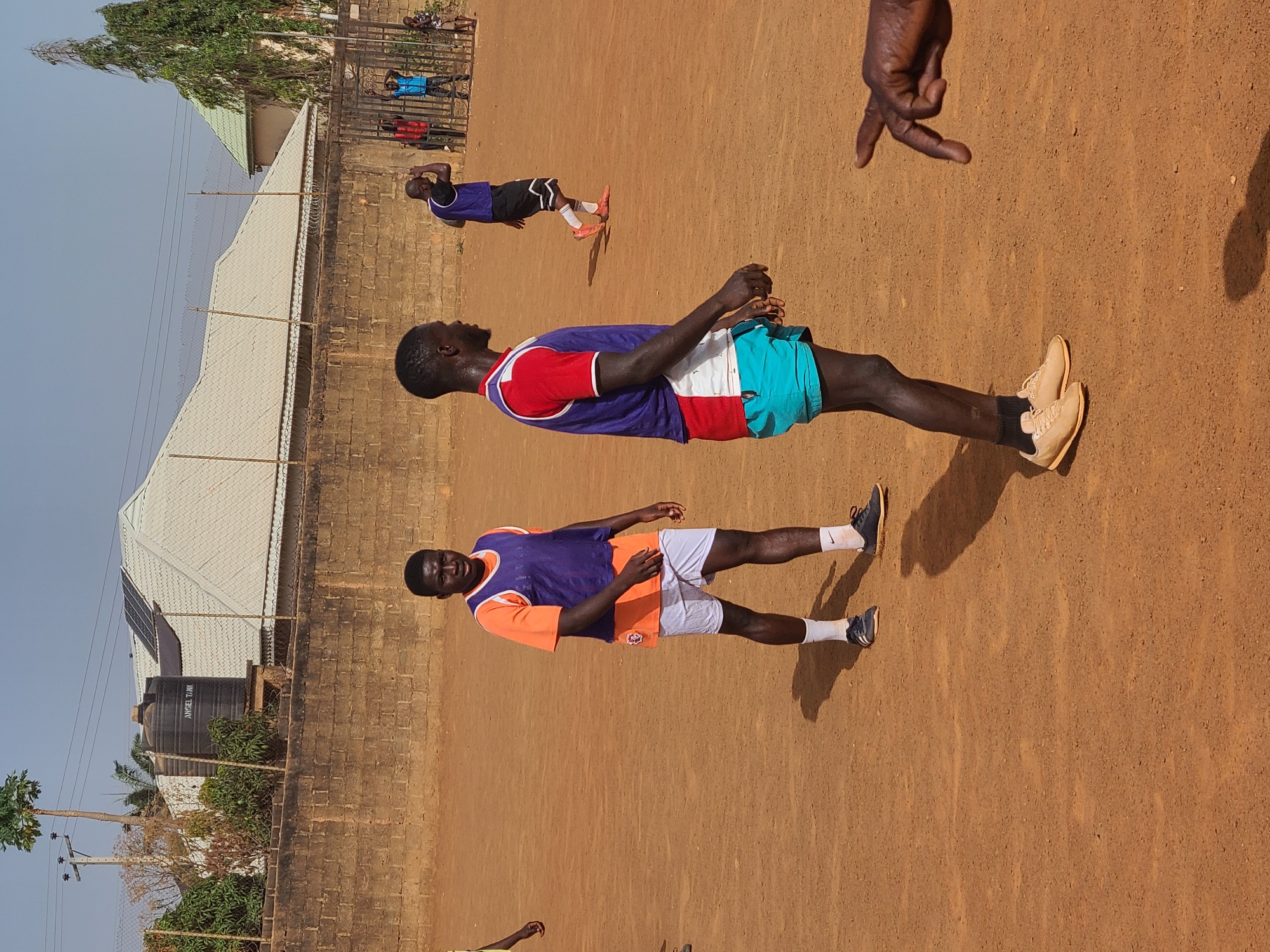 The image shows an outdoor scene in Barakin Lerawi, Nigeria. Three males of African descent are present on a dirt field. One man stands wearing a purple vest with a red neckline, red shoulder stripes, white midsection, and teal shorts. He has tan shoes with black socks. Another man stands nearby, wearing a purple vest with an orange neckline and orange shoulder stripes, and white shorts. He has white socks and dark shoes. In the background, another man lies on the ground wearing a dark shirt and shorts, with orange footwear. A brick wall runs along the edge of the field, with houses and vegetation visible behind it. A water tank and solar panels are visible on a rooftop. The sky is light blue. A dark hand is partially visible in the upper right corner of the image.