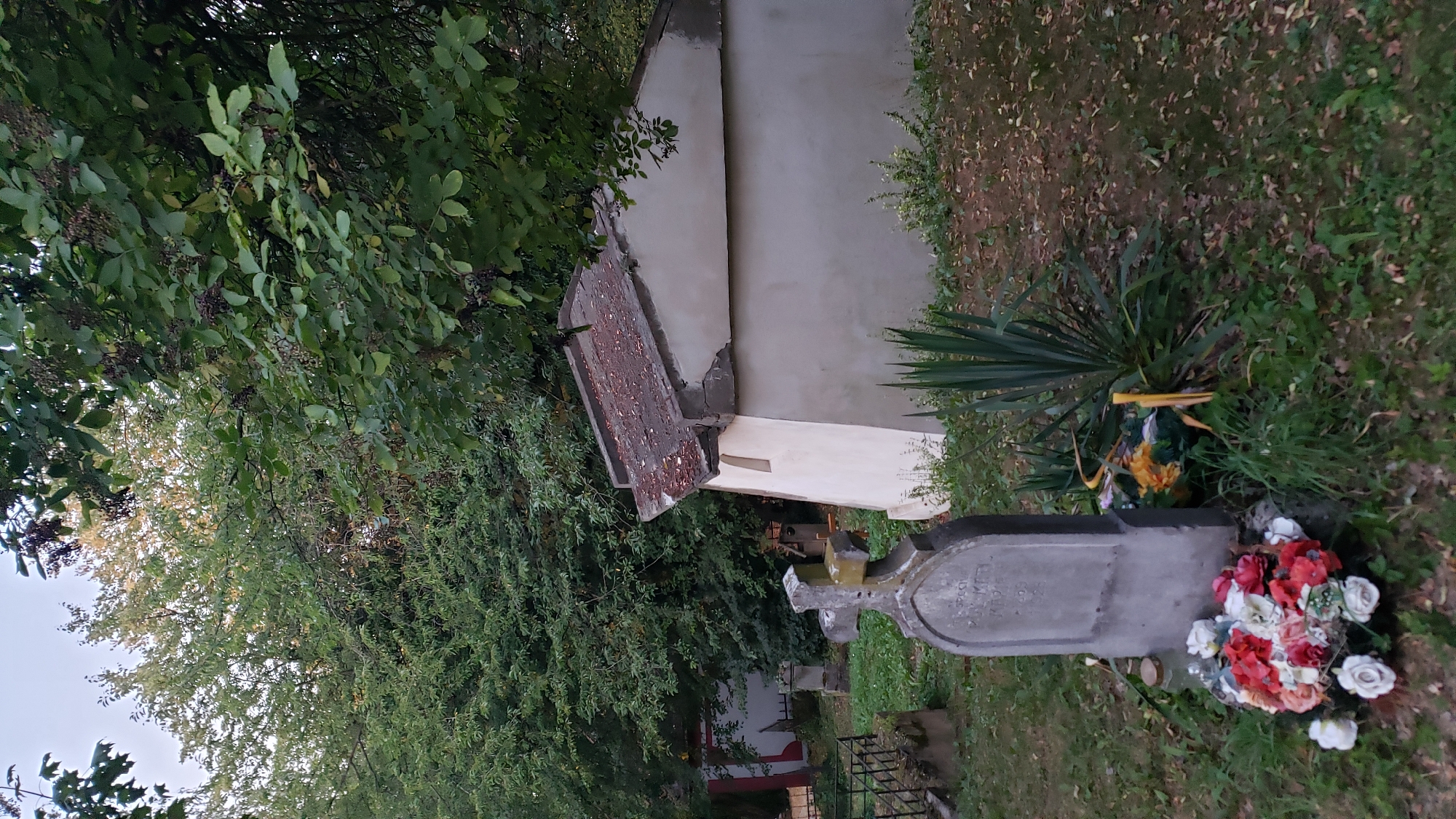This image depicts a cemetery scene, likely in the city of Prešov, Slovakia, though specific identifying markers are absent. In the foreground, a weathered, upright headstone with a cross on top stands prominently. The headstone appears to have inscriptions, partially obscured by time and wear. A floral wreath, composed of red, white, and pink artificial flowers, rests at the base of the headstone. To the right of the headstone, a tall, spiky plant, possibly a Yucca, grows. Behind the headstone and to the right, a small, light-colored building with a sloped, tiled roof is visible. The building has a single, narrow, arched window on its visible side. The overall environment suggests a cemetery with overgrown grass, fallen leaves, and mature trees providing shade. Other gravestones and funerary markers are scattered throughout the background, indicating a densely populated burial ground. The lighting suggests it is late afternoon or early evening. There are no individuals visible in the image.
