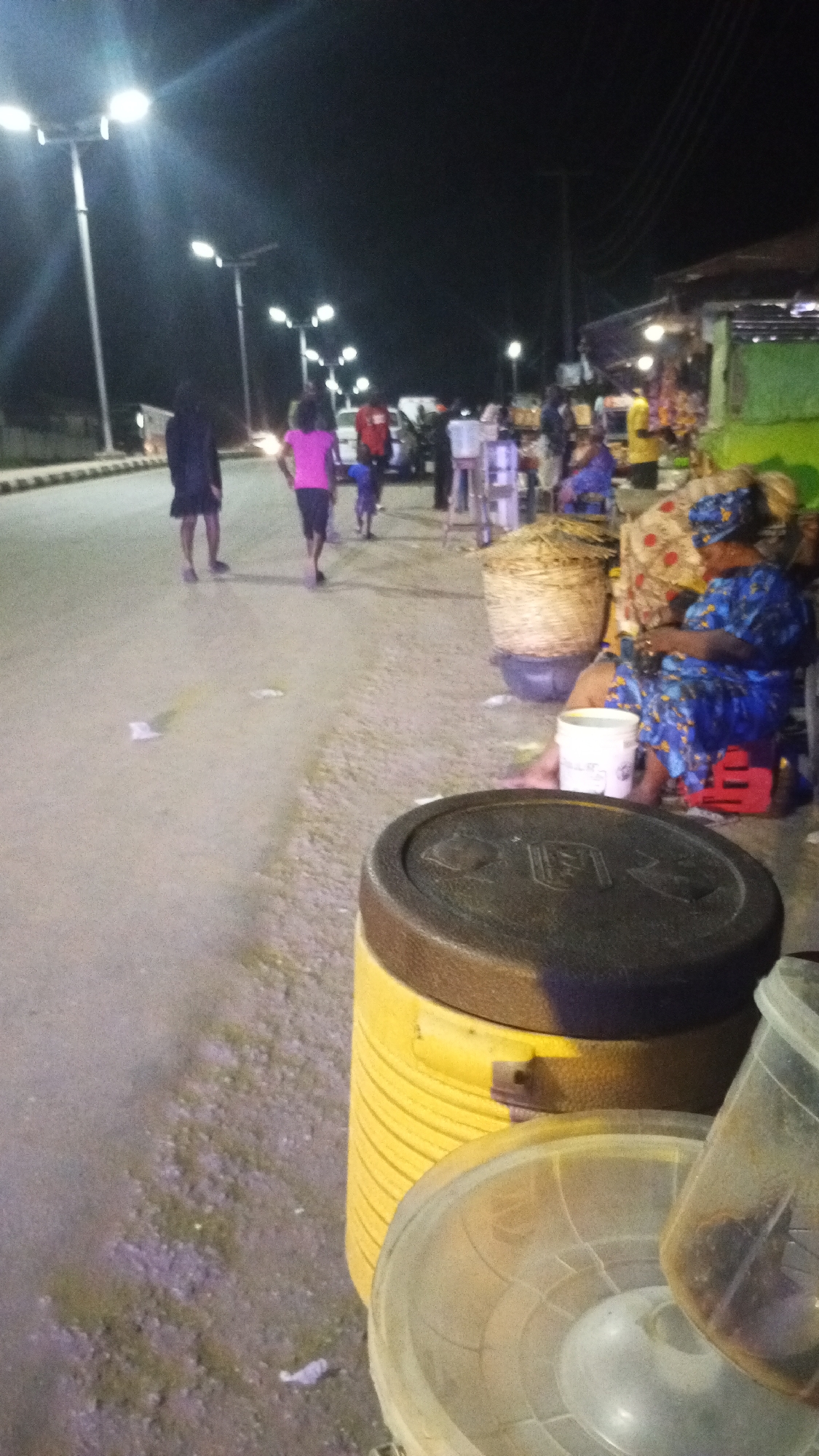 The image shows a street scene at night in Adankolo, Nigeria. Streetlights illuminate the scene. On the left, two people walk along the road. Further down the street, a child follows an adult. A group of people stand near parked vehicles. On the right side of the image, a woman sits on a red plastic stool, near a basket and under a draped cloth. In the foreground, there are yellow and clear containers, possibly for food or storage. Litter is scattered on the ground. There appears to be shops to the right of the image.