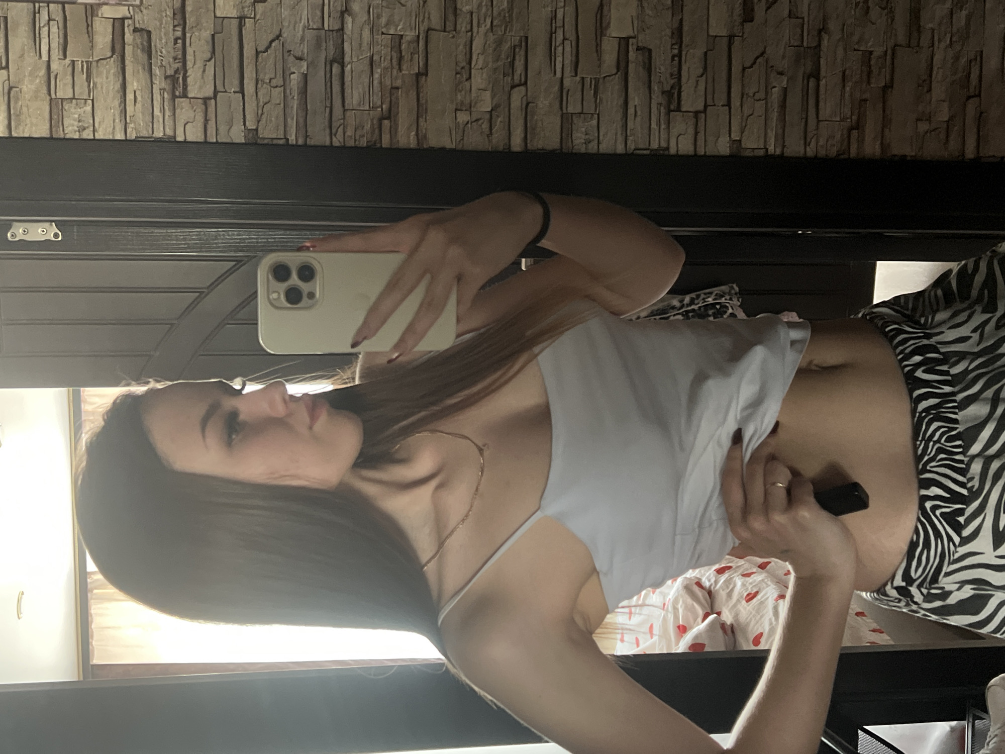 A woman is taking a selfie in what appears to be a bedroom. She is holding a smartphone in her left hand, pointing the camera towards herself. The woman is wearing a white tank top and zebra-print pants. Her hair is long and brown. She is also wearing a bracelet on her left wrist and a gold necklace. With her right hand, she seems to be holding the edge of the top. The background includes a patterned wall and a bed with a patterned bedsheet. The location and city of Noviy, Russia are not discernible from the image.