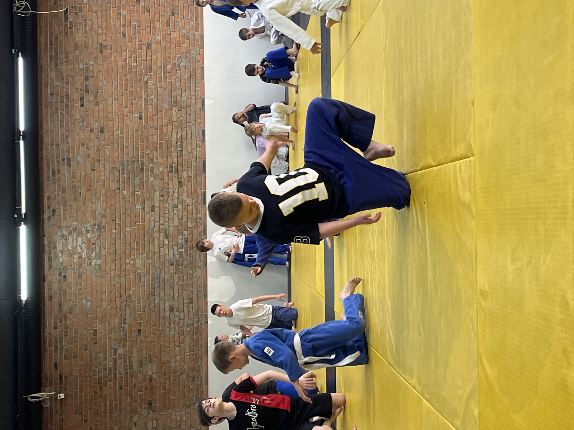 The image depicts a group of children and young teenagers practicing martial arts, likely judo or a similar discipline, on a yellow mat in an indoor training facility. The room has exposed brick walls and fluorescent lighting, suggesting a functional, perhaps urban, setting. Several individuals are dressed in traditional martial arts uniforms, known as gis, predominantly in white and blue.
In the foreground, a boy in a blue gi is kneeling on the mat with a white belt. To his left, another boy in a black t-shirt with a white number "10" and blue pants is in a dynamic pose, appearing to be mid-movement or demonstration. Other participants are scattered across the mat, some kneeling, others sitting, and some standing. Their ages appear to range from approximately 6 to 15 years old.
The backdrop includes a rough brick wall and a white wall section. The lighting is bright and even, illuminating the training area. The overall atmosphere suggests an active training session or class. The location is identified as being in the city of Krasnoyarsk, Russia, which would place this martial arts facility within that specific geographic context.
