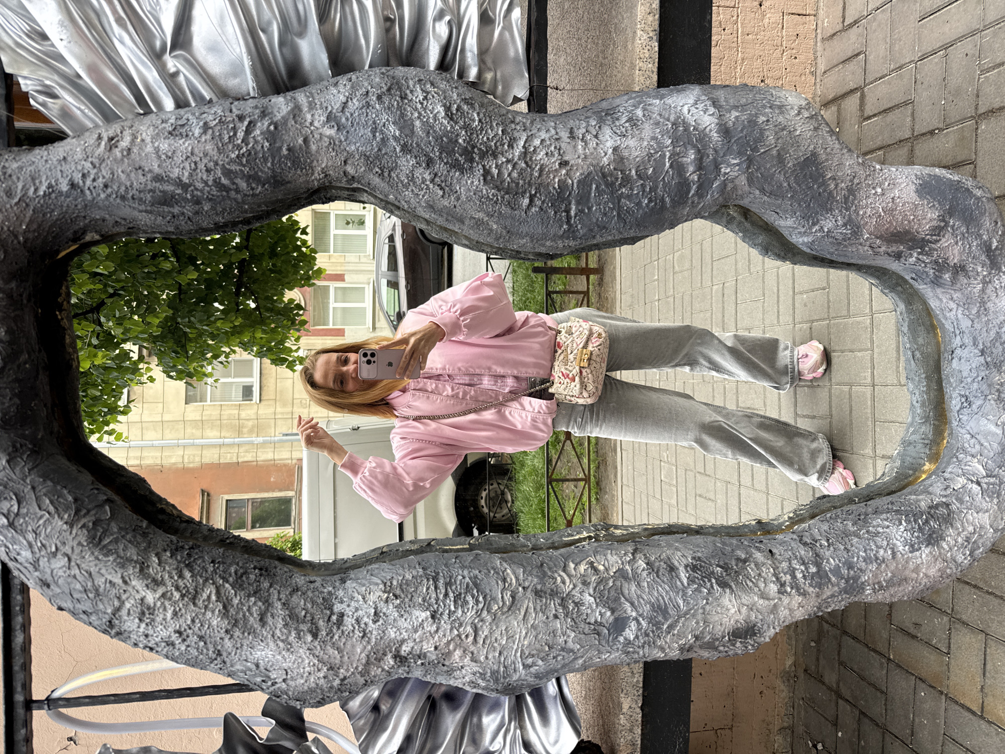 A woman in a pink jacket and gray jeans is taking a selfie in front of a public artwork. The artwork is a large, silver, irregularly shaped frame, with an ornate, textured surface. The frame is positioned on a sidewalk made of gray pavement stones. The reflection of the surrounding environment is visible in the artwork, including a building with windows, green trees, and a white van. The woman holds a phone in her right hand. A patterned purse hangs on her left shoulder. The image appears to be taken outdoors during daylight. The location is in Saint Petersburg, Russia.