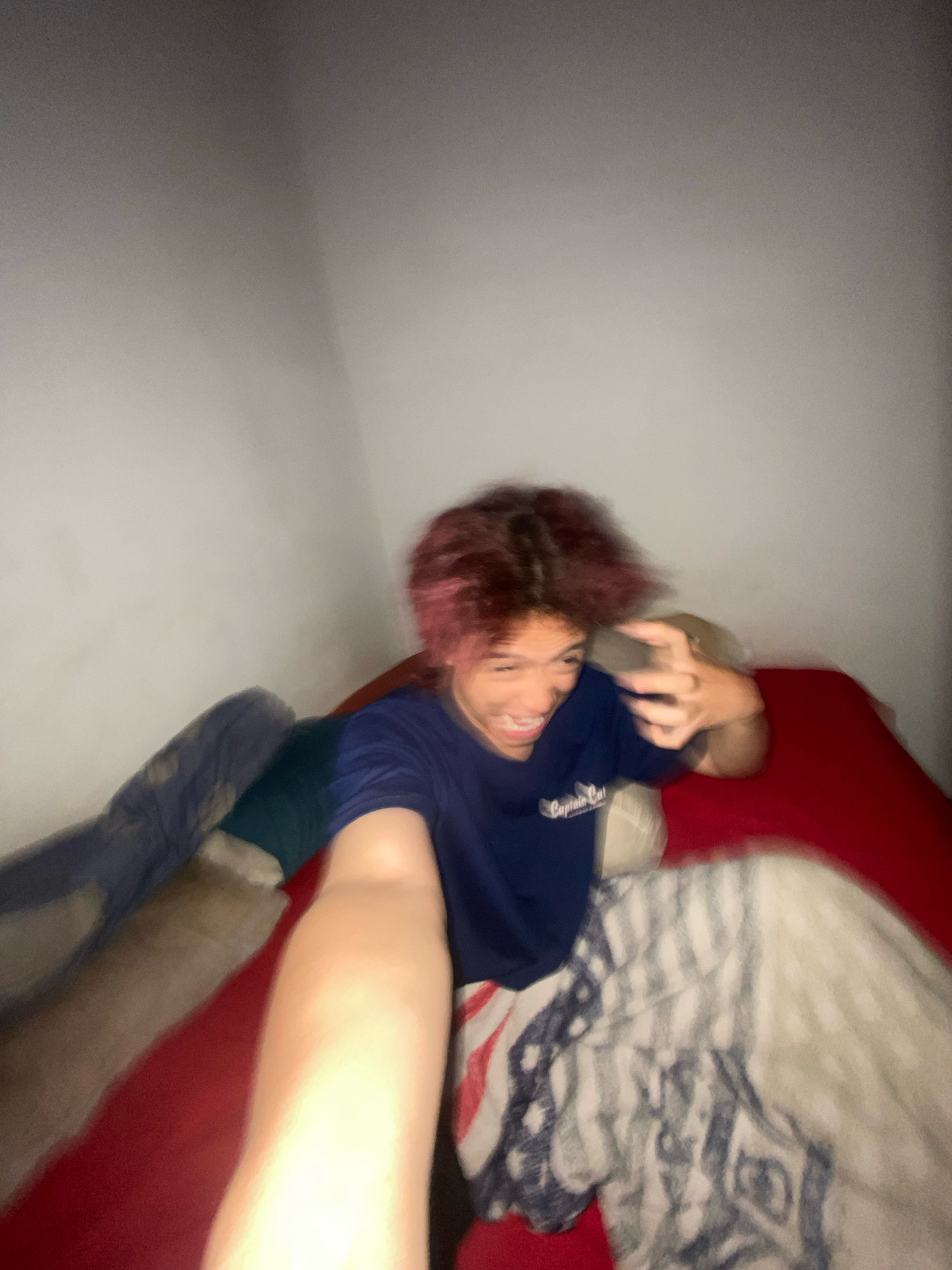 The image depicts a young person with pink-tinted hair, wearing a navy blue t-shirt with a logo, sitting on a bed. They are holding up their right hand, with their fingers splayed. The person appears to be in a bedroom, as indicated by the presence of a bed with red sheets and a blanket with a patterned design that resembles the American flag. The background consists of a plain, white wall. The lighting in the room is dim, and the image is somewhat blurry, suggesting it may have been taken with a mobile phone or in low light conditions. No specific location in the city of Klurak, Indonesia can be determined from the image.