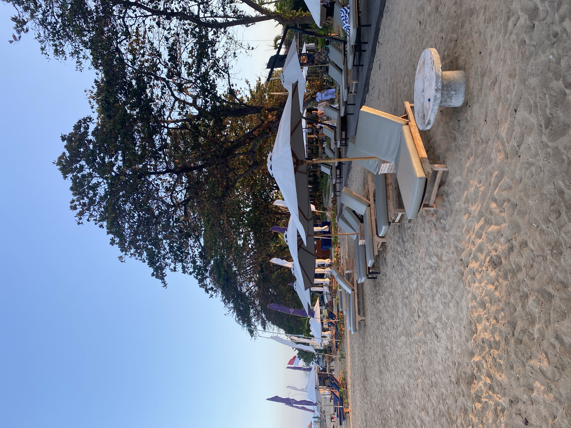 The image depicts a beach scene at what appears to be the sunset in Denpasar, Indonesia. In the foreground, there are several empty lounge chairs made of wood and light gray cushions, arranged in rows on the sandy beach. A low, round concrete table sits in front of one of the lounge chairs. In the middle ground, more lounge chairs and umbrellas are visible, with a wooden boardwalk behind them. Several people are seen walking and interacting near the boardwalk and under the trees in the background. The beach is lined with tall trees and several flags, including what appears to be the Indonesian flag. The sky is clear and light blue, suggesting the late afternoon or early evening. The overall scene conveys a relaxed and serene atmosphere, typical of a beach resort.