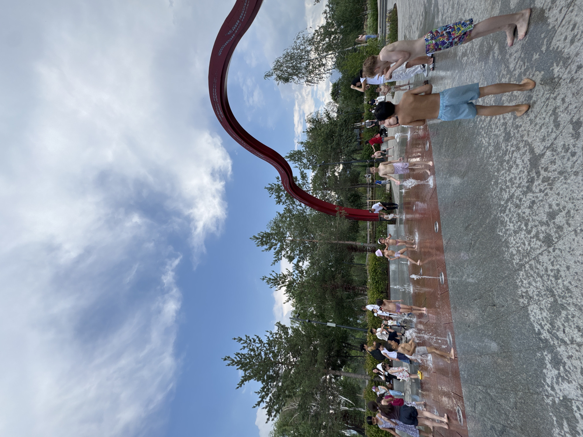 The image depicts an outdoor splash pad area in Almaty, Kazakhstan, likely within a park or recreational space, given the presence of trees and a large red archway structure. Several children and some adults are present, engaged in water play. The ground is wet from the water jets, creating a glistening surface.
In the foreground, two boys are prominently featured. The boy on the left is wearing blue striped shorts and is facing away from the camera. The boy on the right is wearing colorful patterned shorts and is leaning against a wet, grey tiled surface.
Further into the scene, more children are visible playing in the water. Some are wearing swimsuits, while others appear to be in casual summer clothing. Adults are scattered throughout the area, observing or participating.
Behind the splash pad, a dense line of coniferous trees forms a backdrop, with some deciduous trees also present. The sky above is partly cloudy, with patches of blue sky visible. The red archway, a prominent architectural feature, spans across the upper portion of the image, its curved design contrasting with the natural elements. The overall impression is a lively, summer day scene with people enjoying a water attraction.