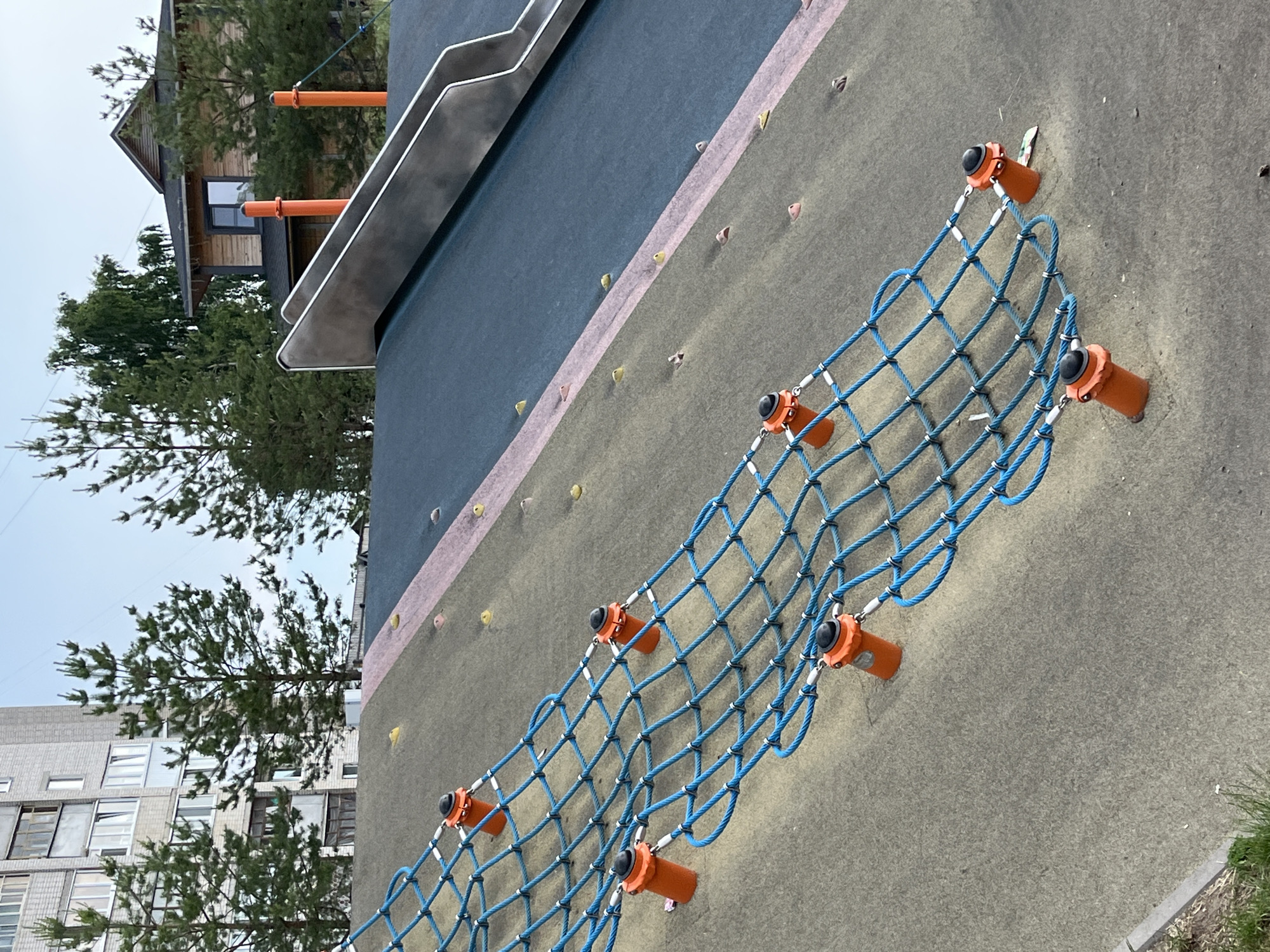 The image depicts an outdoor playground in Zelenodol'sk, Russia. The foreground features a climbing net made of blue ropes stretched across a sloped, textured surface. The net is secured by several orange posts topped with black caps. Small, colored climbing holds are embedded in the sloped surface, some pink and some yellow. A portion of a dark blue slide is visible in the upper right quadrant of the image, with metal rails. In the background, a tall, grey apartment building stands to the left, partially obscured by evergreen trees. To the right of the apartment building, a wooden house with a dark roof is visible, also surrounded by trees. The sky is overcast and a pale blue.