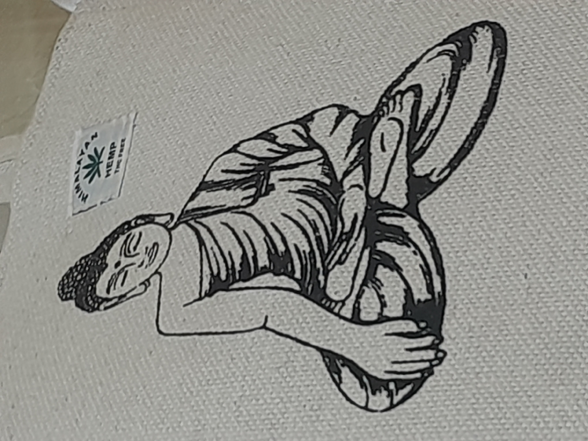 An illustration of a seated Buddha is printed in black ink on a light-colored, textured fabric, likely a tote bag or textile. The Buddha is depicted in a cross-legged pose, with one hand resting on the knee and the other partially visible. The figure has a serene expression, with closed eyes and a simple robe draped around the shoulders. Above the Buddha's head, to the upper right, is a small rectangular label with an image of a hemp leaf and text that reads "HIMALAYAS HEMP THE FREE." The background is a uniform, textured surface. The image appears to be a close-up shot of the fabric. The location is not identifiable from the image itself.