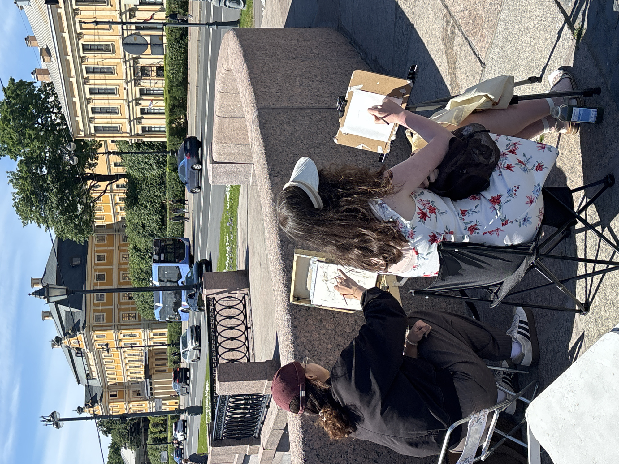 Two individuals are engaged in plein air painting by a stone embankment overlooking a street in Saint Petersburg, Russia, with buildings and trees in the background. The person on the left, wearing a burgundy baseball cap, glasses, a black jacket, and dark striped pants, is seated on a white folding chair and is actively drawing on a portable easel. They are wearing white athletic shoes with black stripes. To the right, another individual, wearing a white visor and a floral print dress, is also seated on a black folding chair and is drawing on a portable easel. They have a dark bag slung over their shoulder and are wearing sandals with white socks. A green bottle is placed on the ground near their feet. The embankment is made of granite blocks, and a decorative black metal fence with oval patterns is visible behind the artists. Across the street, a blue bus and several cars are moving, and large, light-yellow buildings with ornate architecture line the street. The sky is clear and blue, indicating a sunny day. The scene appears to be along one of the many canals or rivers in Saint Petersburg, given the presence of the embankment and the urban cityscape.