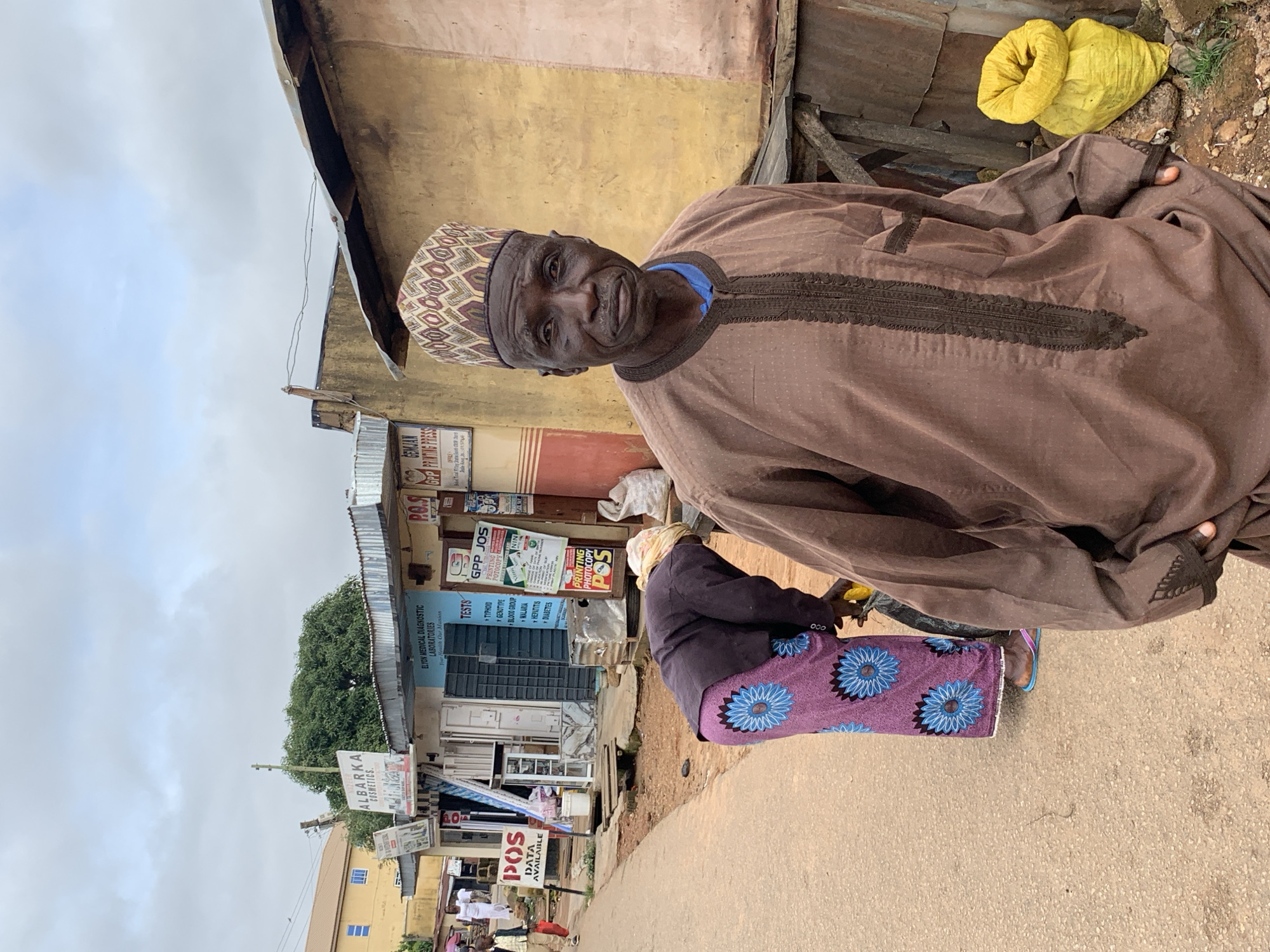 In Jos, Nigeria, a man in a traditional brown robe and a patterned cap stands in the foreground, looking directly at the camera. Behind him, a woman in a dark jacket and a purple skirt with blue floral patterns is bending down. In the background, several small shops line the street, with signs indicating businesses such as "ALBARKA COSMETICS," "ELTON MEDICAL DIAGNOSTIC LABORATORIES," and "GPP PRINTING PRESS." The street is paved with rough asphalt, and the buildings are constructed with corrugated metal and faded paint. Two yellow sacks are piled near a wooden structure to the right of the frame. The sky is overcast with clouds.