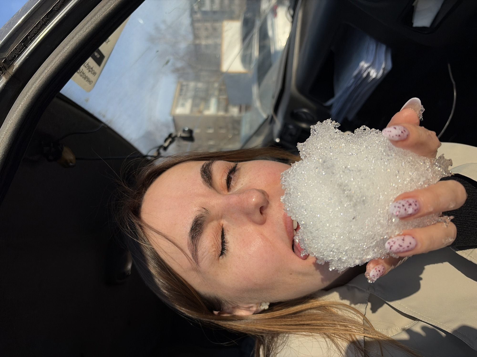 A young woman is holding a handful of snow to her mouth inside a car. She has her eyes closed and appears to be eating the snow. The woman has light skin, brown hair with blonde highlights, and manicured nails with a white base and pink spots. The car's interior is visible, with a window in the background showing a blurred view of buildings, a potential location in the city of Izhevsk, Russia. The building looks very distant and out of focus. The sunlight is shining in. The woman is wearing a light beige or white jacket.