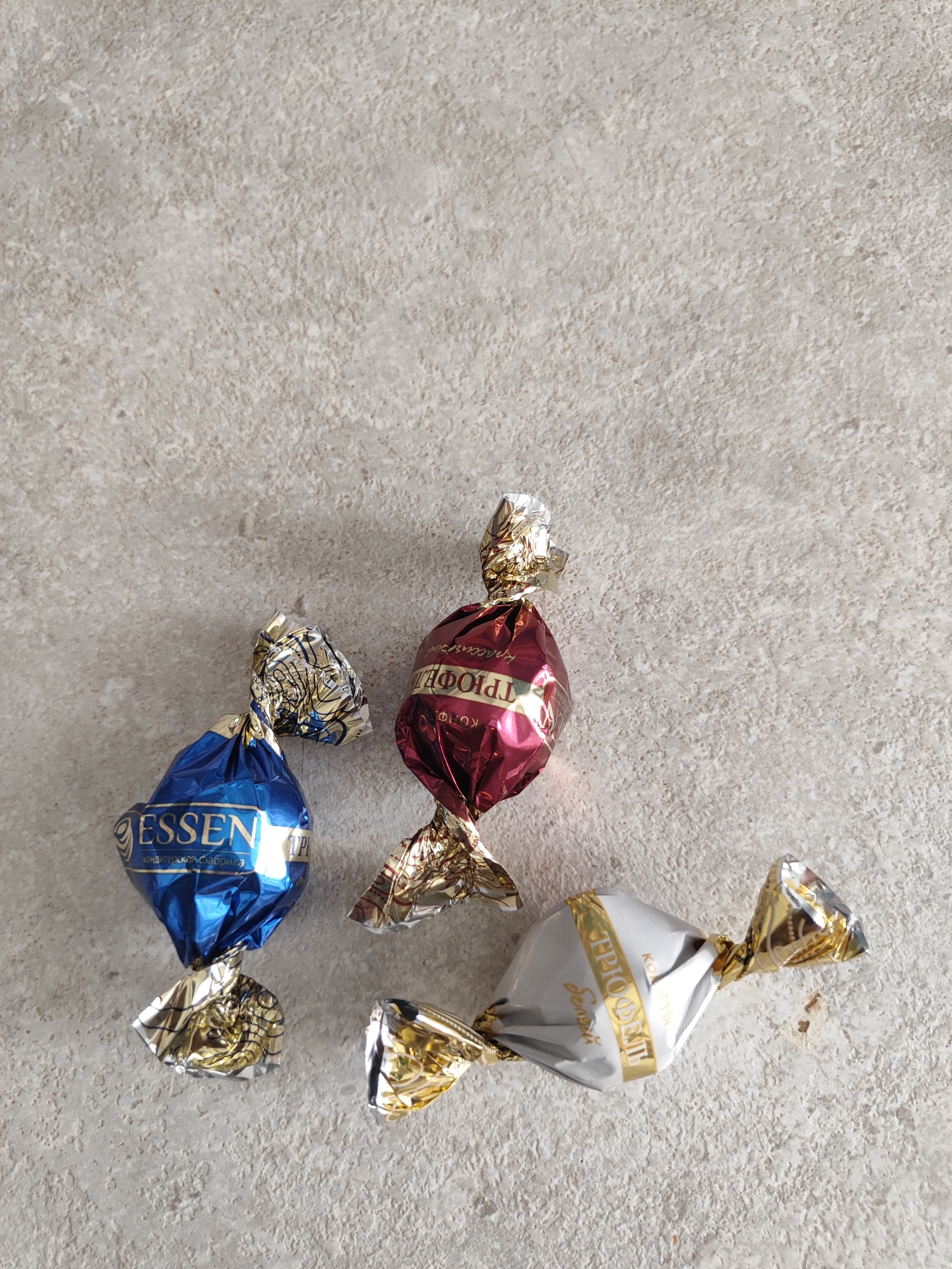 Three individually wrapped candies are arranged on a speckled gray surface. From left to right, the first candy is wrapped in blue foil with gold accents and text that reads "ESSEN" in large gold letters. The second candy is wrapped in dark red foil with gold accents and text that appears to be in Cyrillic. The third candy is wrapped in silver foil with gold accents and text that reads "TRIO DONT" in gold letters. The background appears to be a neutral-toned textured surface, possibly a countertop or floor. The lighting suggests the image was taken indoors.