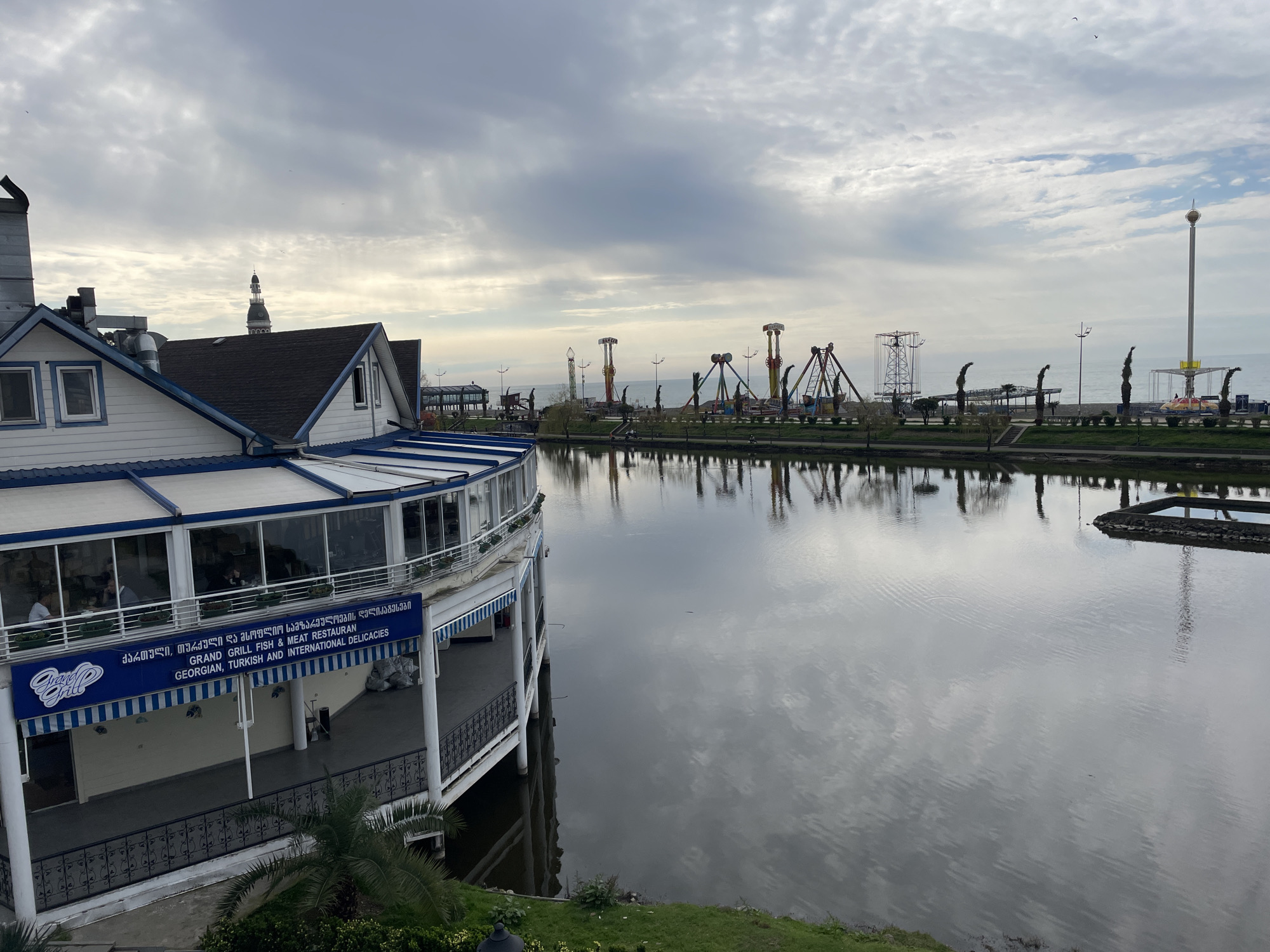 The image shows a restaurant on the edge of a body of water in Batumi, Georgia, with an amusement park in the background. The restaurant, identified by a sign that reads "Grand Grill Fish & Meat Restaurant," features Georgian, Turkish, and international delicacies. The building is painted white with blue trim and has a glass-enclosed dining area. The water reflects the cloudy sky above. In the distance, a range of amusement park rides, including swings and towers, are visible along the waterfront. The scene is captured from a slightly elevated position.
