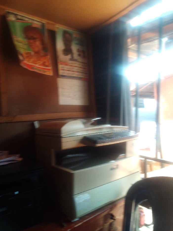 The image shows a beige multi-function printer sitting on a wooden table. The printer is turned on and ready to use. The scene is located in Auchi, Nigeria. The printer is likely in a shop or office. The background shows a wall with a bulletin board. The bulletin board has two political posters on it. There is a black chair with a white plastic seat in the foreground of the image. The window is covered by black curtains. The window looks out onto a street. The scene is lit by bright sunlight coming from the window. The room is small and cluttered. The image was taken from a low angle.