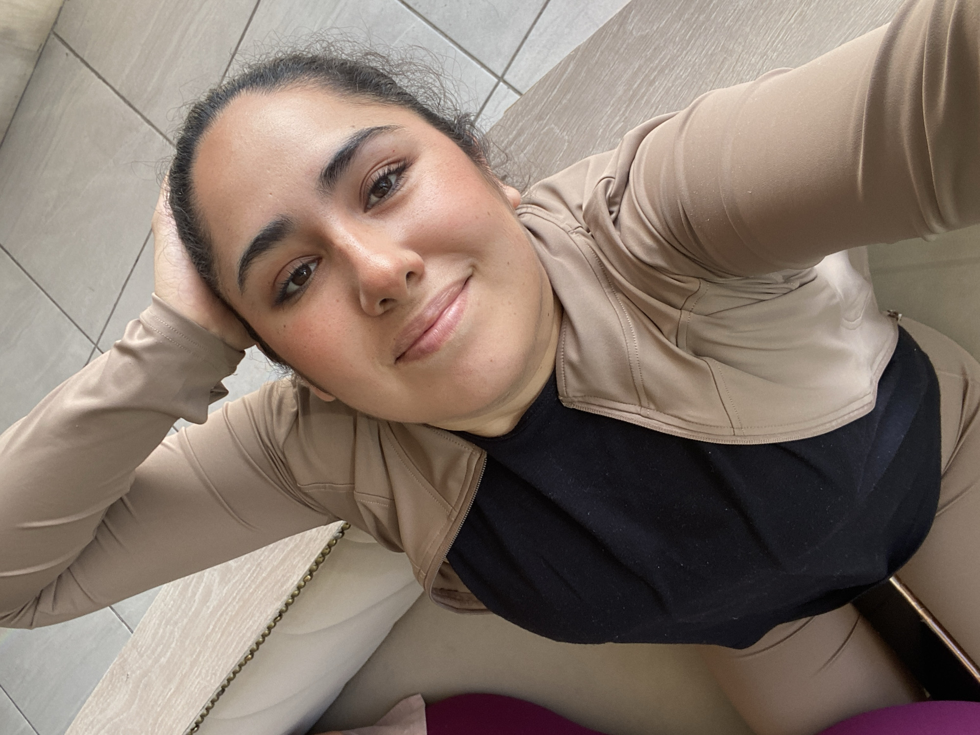 A person with medium brown skin and dark hair is posing for a selfie indoors. They are wearing a black shirt under a light brown jacket. The person has a slight smile and is resting their head on their hand. The background includes tiled flooring, a light-colored chair, and a glimpse of a purple item. The location is most likely indoors, but without further information, the city of Puebla, Mexico, cannot be specifically determined.