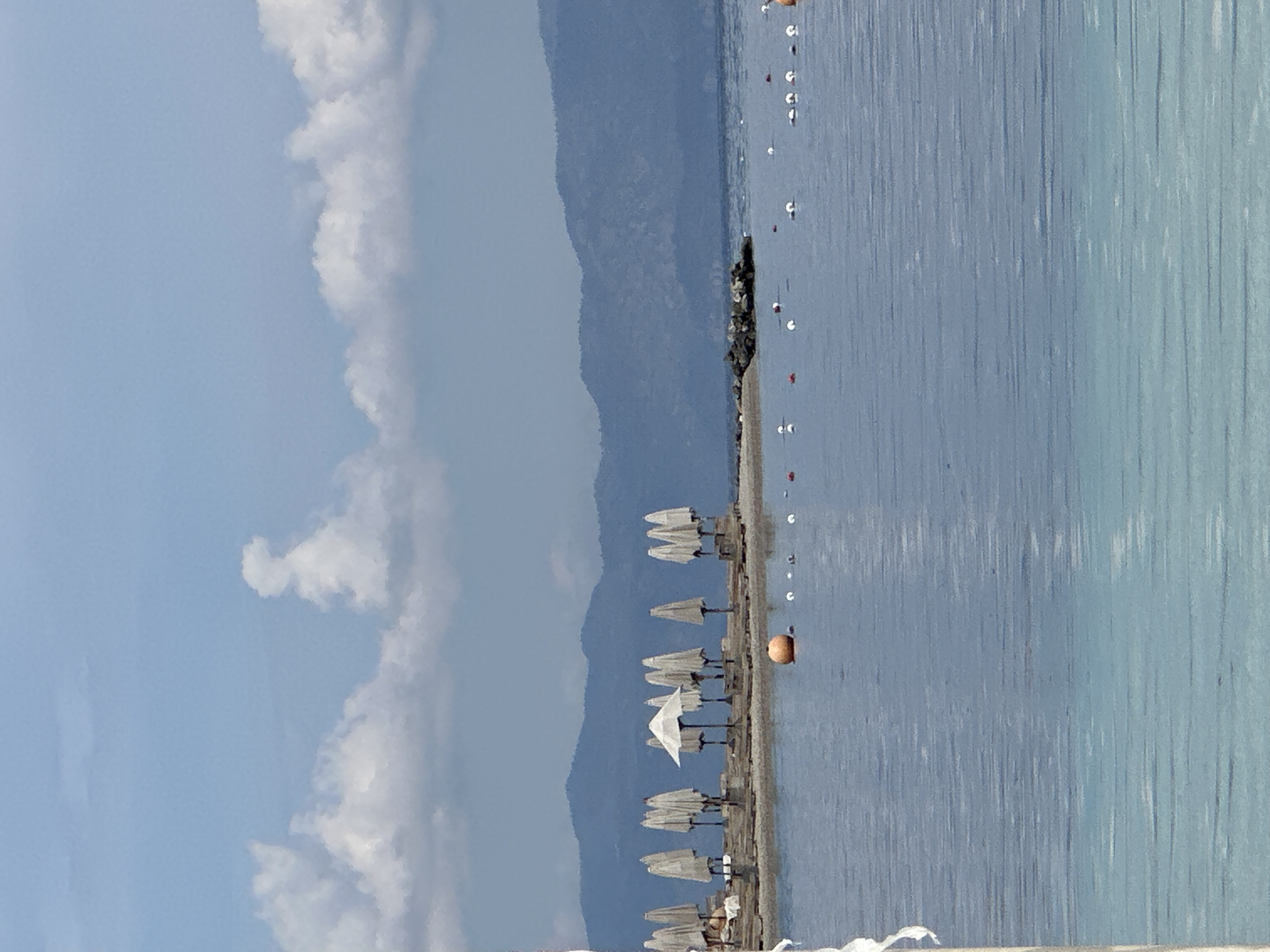 The image depicts a beach in Fethiye, Turkey. The scene is composed of a coastline, a body of water, and a sky with clouds. The coastline has a row of closed white beach umbrellas, and the water has several buoys. A mountain range is visible in the background. The sky is mostly blue with scattered white clouds.