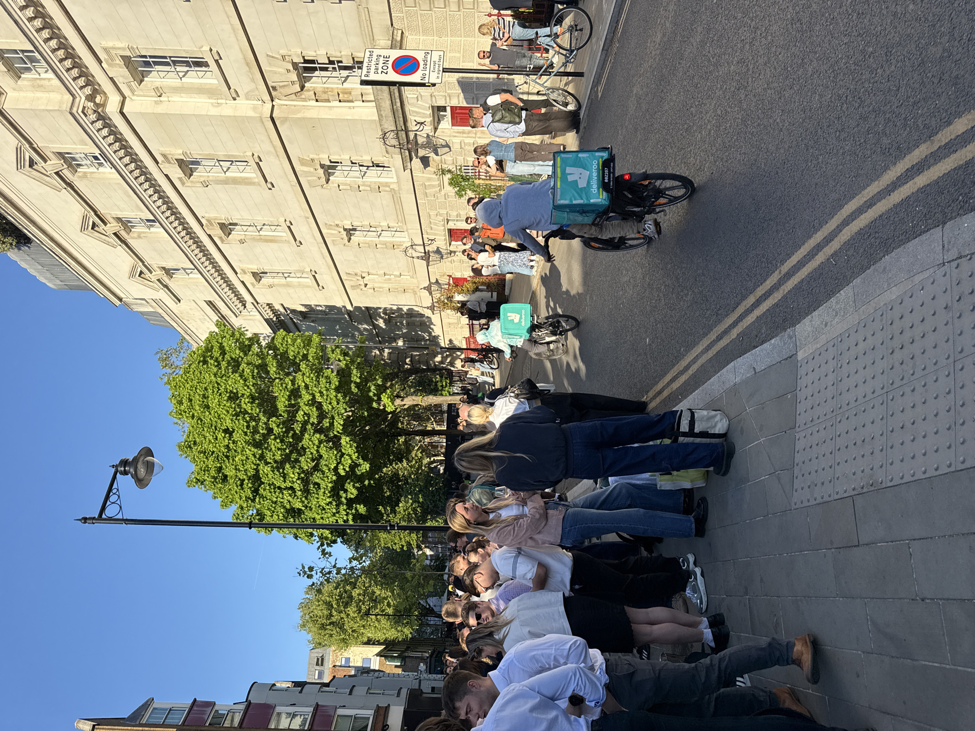 The image, taken in London, United Kingdom, depicts a lively urban street scene on a clear, sunny day, likely during late morning or afternoon judging by the bright light and distinct shadows.
In the foreground, a long queue of diverse individuals, primarily young adults, stands along a paved sidewalk. Most are seen from behind, suggesting they are waiting patiently for an event, establishment, or service. To the left, a large tree with vibrant green foliage and a classic black lamppost stand out against a clear blue sky.
The middle ground features a street with asphalt paving and double yellow lines. Several people are visible, notably two bicycle delivery riders wearing blue and carrying distinctive teal Deliveroo thermal bags. One rider is actively cycling away from the camera. Other pedestrians are scattered along the sidewalk.
To the right, a grand, multi-story building constructed of light-colored stone or concrete dominates the background. It features numerous windows and intricate architectural detailing. Several people are gathered on the sidewalk in front of this building, some near red doorways.
Notable visible text includes the "Deliveroo" logo on the delivery bags, with one bag showing the number "882257." A street sign clearly states "Restricted parking zone" and "NO loading except in signed bays." The overall scene captures a typical, bustling city environment.