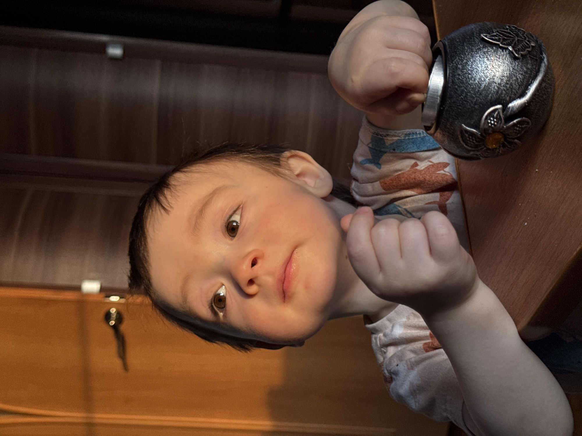 A child with dark hair and brown eyes is reaching for a metallic, spherical object with floral decorations on top of a wooden table. The child is wearing a patterned, long-sleeved shirt. A wooden cabinet with a key is visible in the background. There is no evidence to confirm that the location is Izhevsk, Russia.