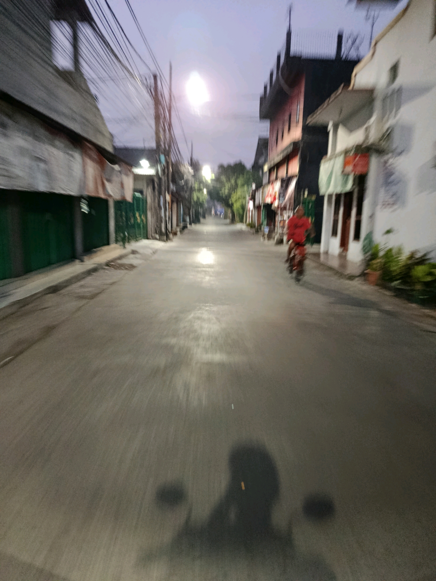 The image depicts a street scene at dusk, likely in Bekasi, Indonesia, characterized by a blend of residential and commercial buildings lining a paved road. The lighting suggests it is either early morning or late evening.
In the foreground, the shadow of a person on a motorcycle is visible on the road surface, indicating the perspective is from the rider. The road itself is asphalt and appears wet in places, reflecting the ambient light.
Further down the street, a person wearing a bright red shirt is riding a motorcycle towards the viewer. The person is blurred due to motion.
On either side of the road, buildings of varying architectural styles are present. On the left, there are structures with corrugated metal awnings and green shuttered doorways. On the right, a white building with balconies and windows is visible, alongside other darker buildings with multiple stories. Utility wires are strung overhead, a common feature in urban Indonesian landscapes.
The sky is a pale purple-gray, with a bright, diffused light source high in the sky, possibly the moon or a light fixture, contributing to the dim illumination of the scene. Trees and foliage are visible in the distance, suggesting the presence of greenery within the urban environment. The overall impression is that of a quiet, possibly residential or mixed-use, street in an Indonesian city during the transitional light of dawn or twilight.