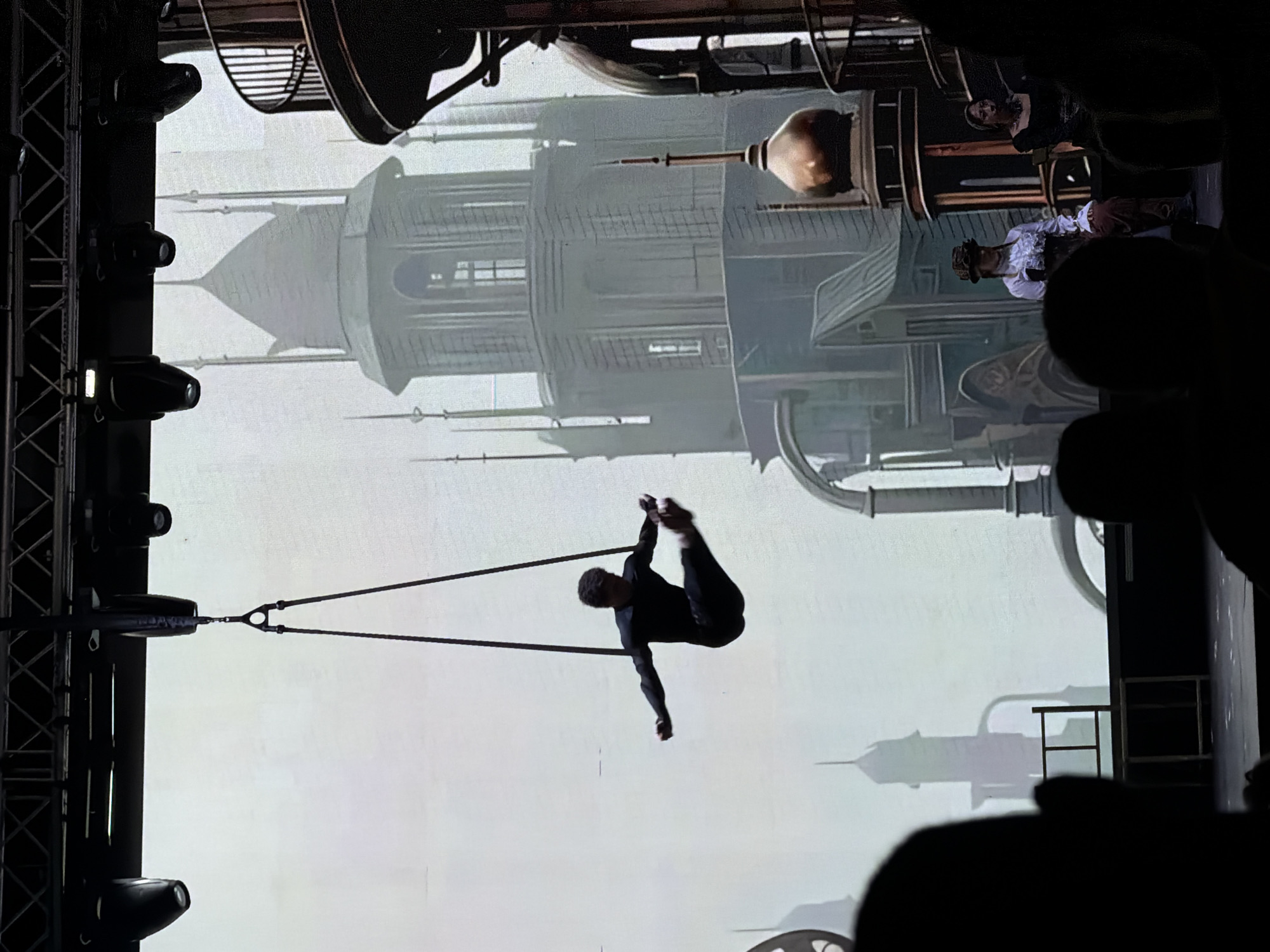 An aerial performer in a black outfit is suspended mid-air from two ropes, performing a pose with their legs extended. The background features a projected digital image of a fantastical, steampunk-inspired cityscape with tall, intricate buildings and spires. In the foreground, two individuals are seated, partially silhouetted. One is wearing a white ruffled shirt, brown pants, and a hat, resembling a steampunk costume. The other is wearing a dark top. The scene appears to be a performance or show. This image was taken in the city of Manavgat, Turkey.