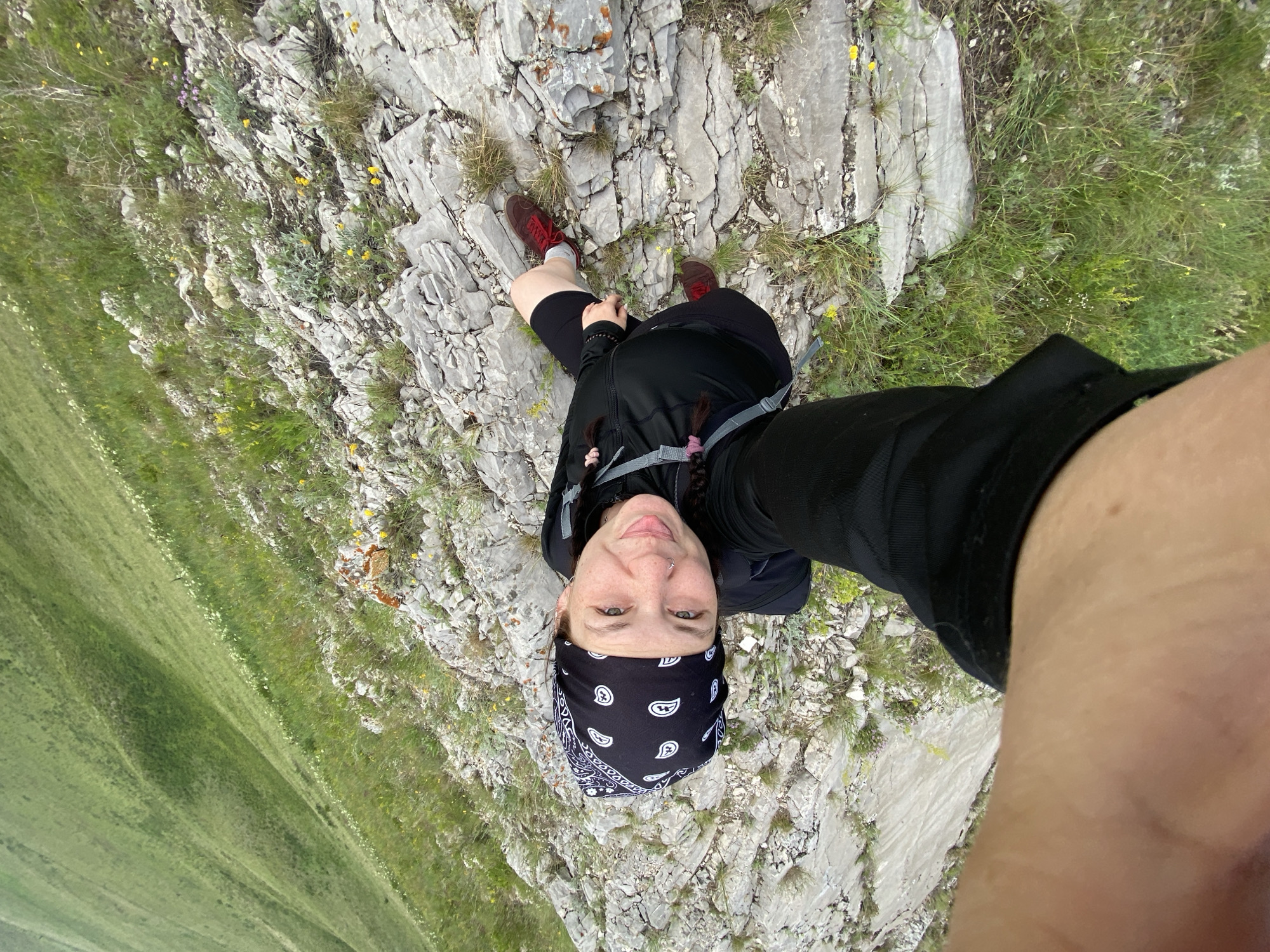 A woman with a bandana and backpack poses for a selfie on a rocky, grassy slope. She is looking at the camera with a smile and her tongue sticking out slightly. Her right leg is extended toward the camera, with a portion of her arm visible in the foreground. The background features a lush green hillside with scattered rocks and small yellow flowers. The location is identified as Verkhnyaya Chernorechka, Russia.