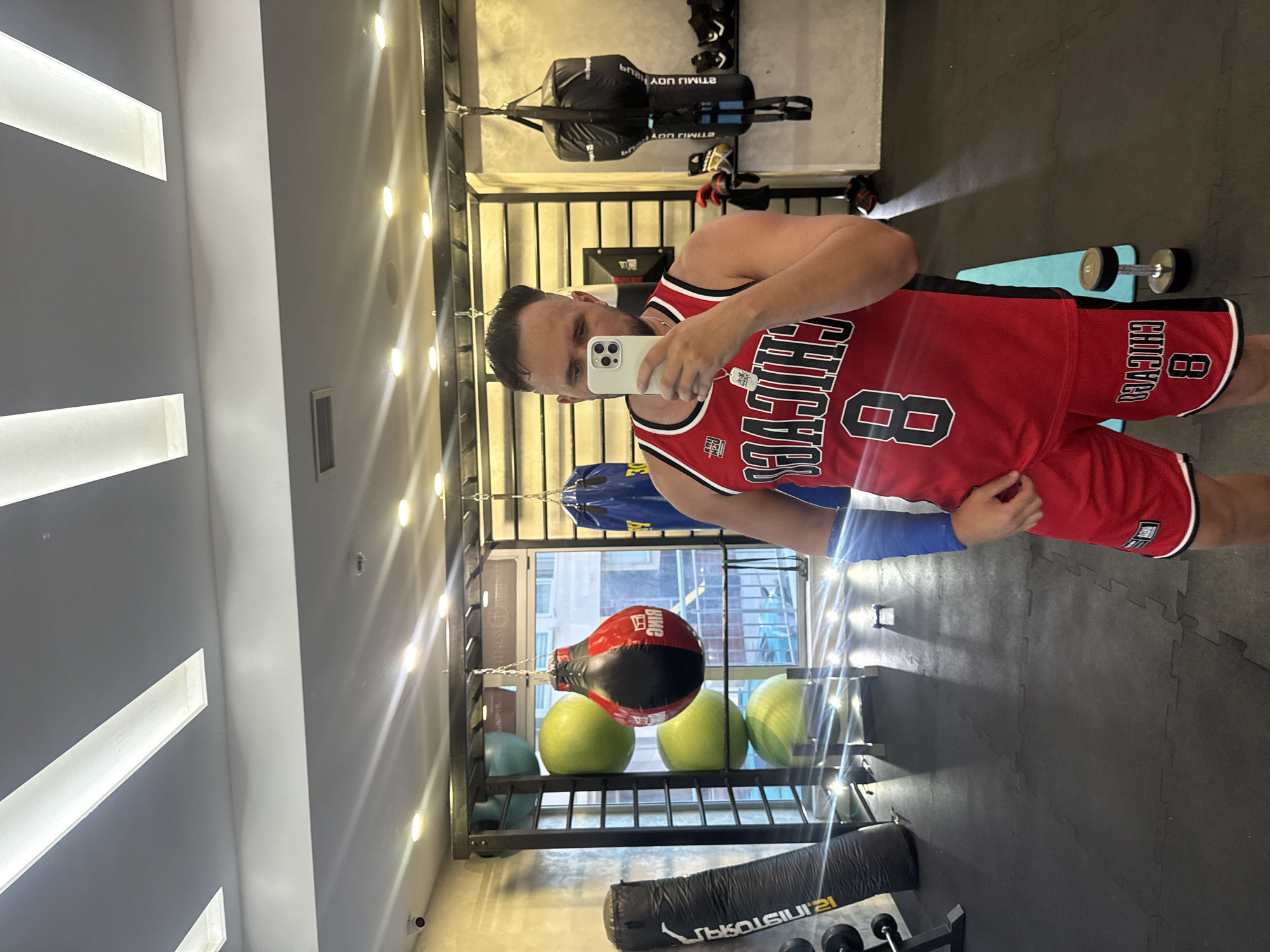 A man is taking a mirror selfie in a gym, wearing a red basketball jersey and shorts with "CHICAGO" and the number "8" printed on them. He has a blue band on his left forearm. The gym is equipped with various workout equipment, including punching bags, medicine balls, and dumbbells. The location is identified as Budva, Montenegro.