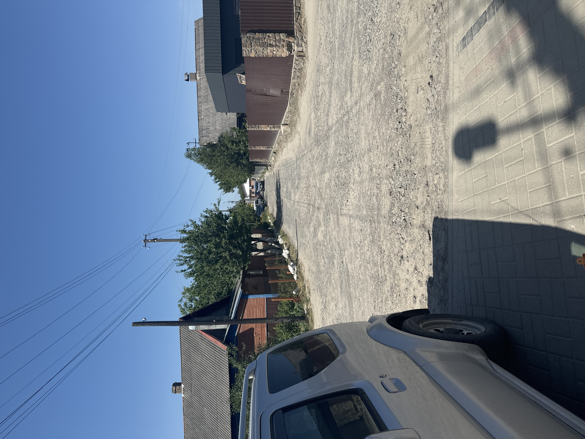 A clear, bright day in Sarny, Ukraine, is captured from a slightly elevated, rotated perspective. The scene looks down a dusty, gravel-lined road bordered by houses and fences on the left and a paved sidewalk on the right. A light-colored, modern SUV is parked close to the camera, its side and spare tire visible.
The road is flanked by a mix of buildings. On the left, closer to the camera, are houses with corrugated metal roofs and wooden siding, interspersed with trees. Further down the road, more houses and fences are visible, along with utility poles and power lines stretching across the bright blue sky. On the right side of the road, the sidewalk is made of interlocking grey bricks. A distinct shadow of what appears to be a lamppost or similar structure falls across the paved surface.
The overall impression is of a sunny, ordinary day in a residential area. There are no people visible, and the focus is on the environment and the parked vehicle. The quality of light suggests it is likely midday.