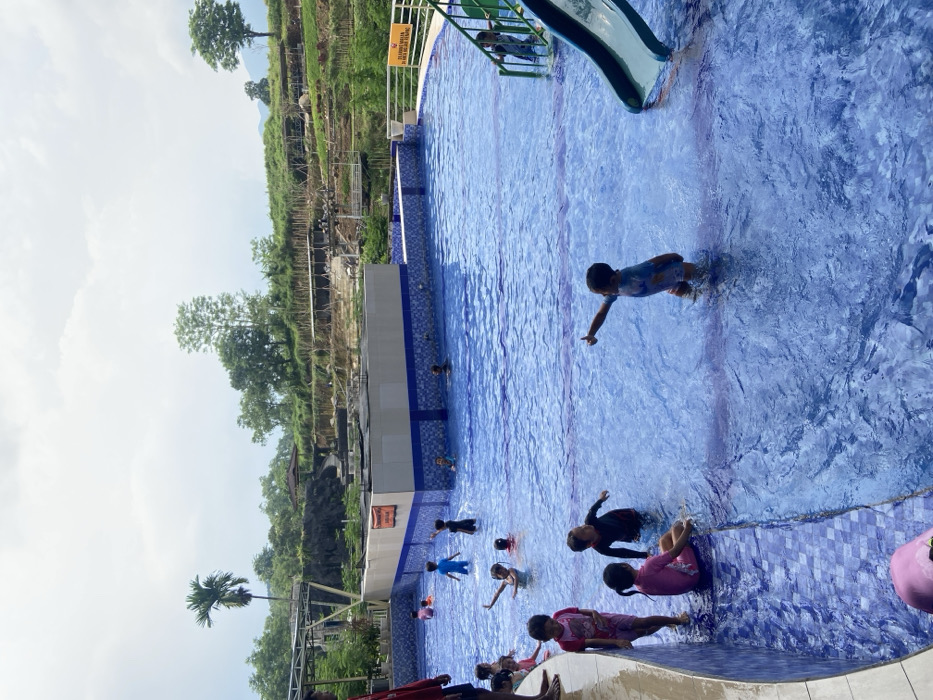 A vibrant outdoor swimming pool teems with children enjoying the water. A blue water slide sits partially visible in the pool's corner. The pool's tiling features a mottled blue pattern. Terraced hillsides surround the pool area, showcasing greenery and what appears to be some sort of construction or support structure. The sky is overcast, but the scene is brightly lit.
