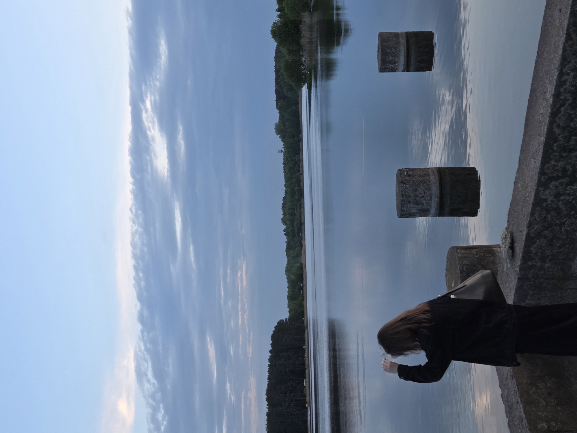 The image shows a woman in a black coat standing on a concrete structure, potentially a pier or dam, overlooking a body of water. The water reflects the cloudy sky above. In the water are two concrete pillars. The woman appears to be holding something in her hand, possibly looking at it closely. The location is Unknown, Unknown. The time of day seems to be dusk or twilight, given the light and sky conditions.